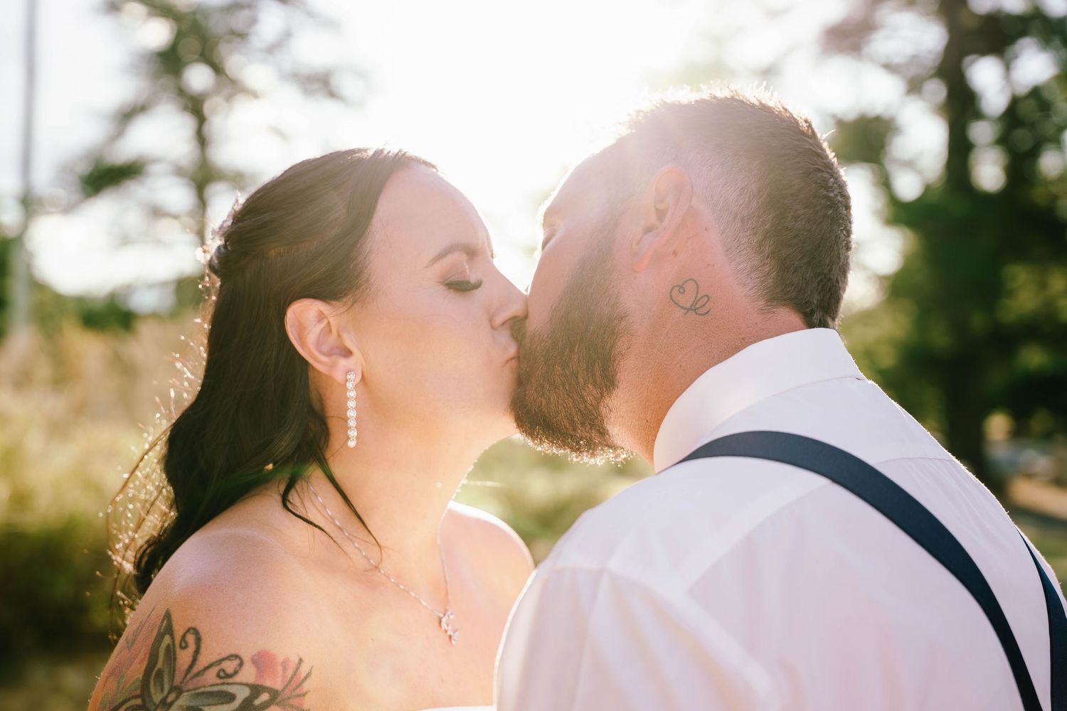 A bride and groom share a romantic kiss outdoors, with golden sunlight glowing behind them on their wedding day.