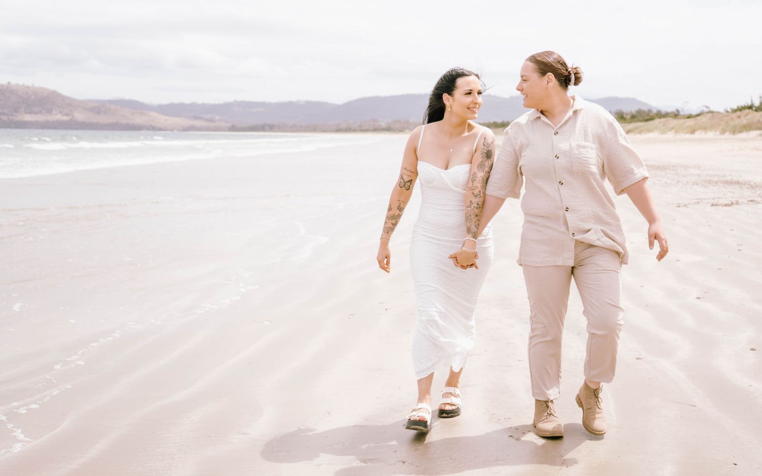 A romantic couple in light beige and white outfits walking together on a sandy beach during sunset.