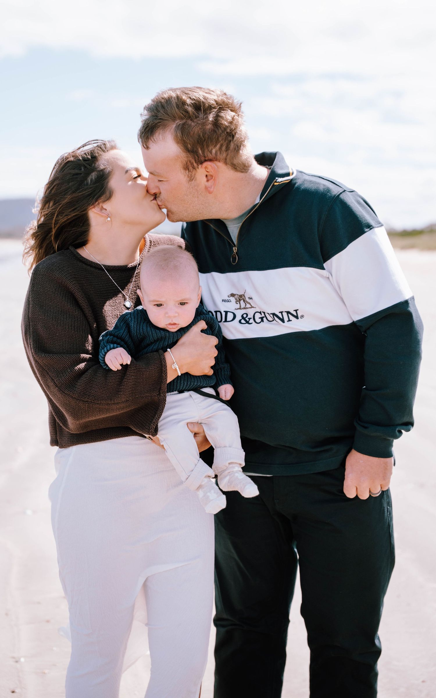 A couple shares a romantic kiss on a beach while holding their baby in coordinating black and white outfits.