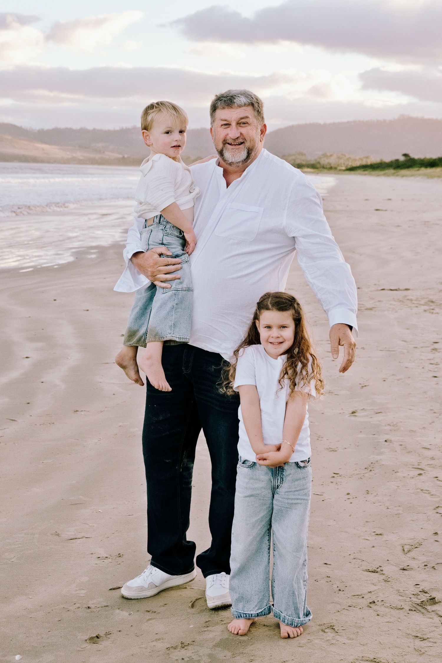 Man in white shirt holds toddler while older girl stands beside him on a sandy beach at sunset.