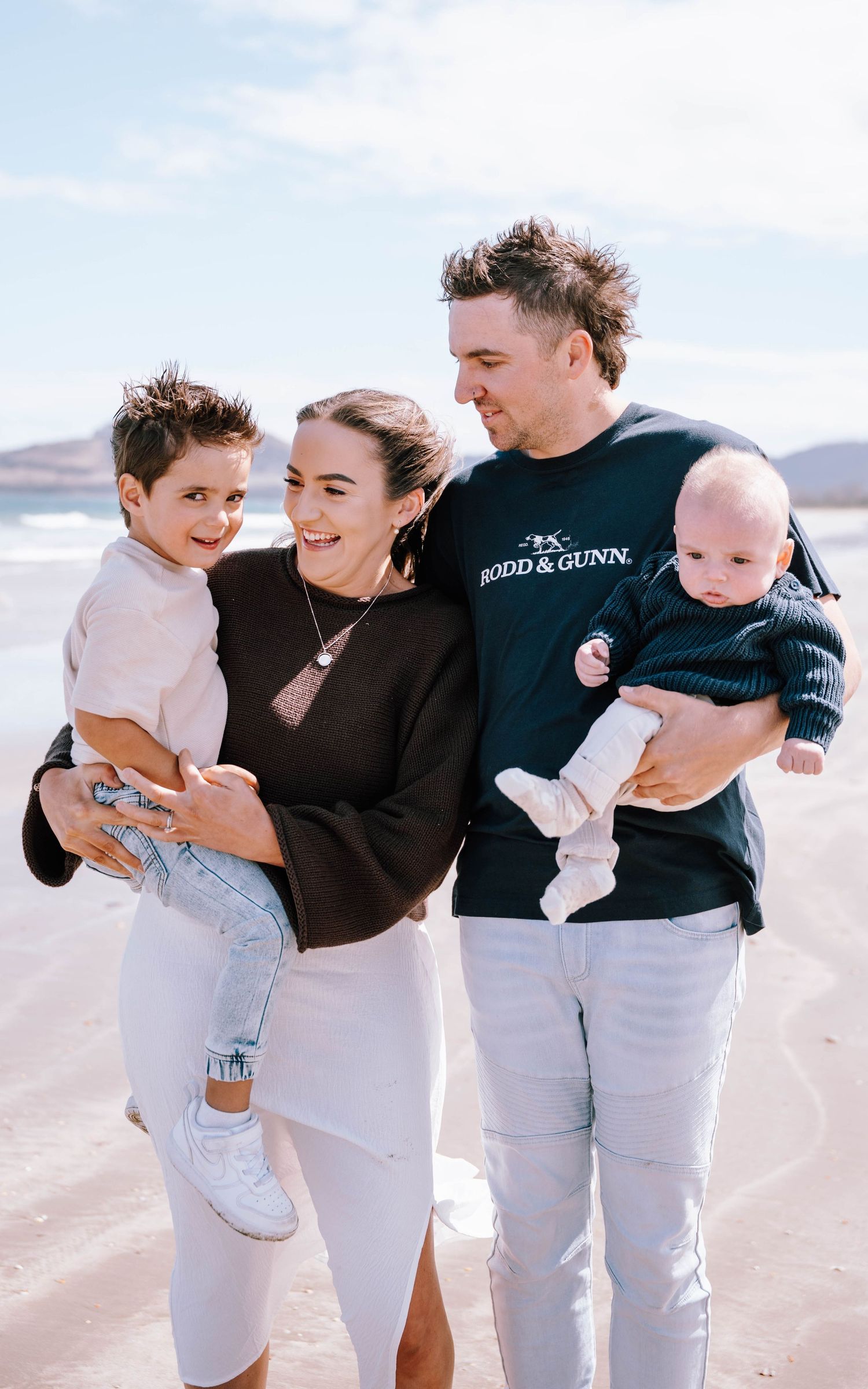 Family wearing matching black tops and light colored bottoms embraces on a sunny beach.