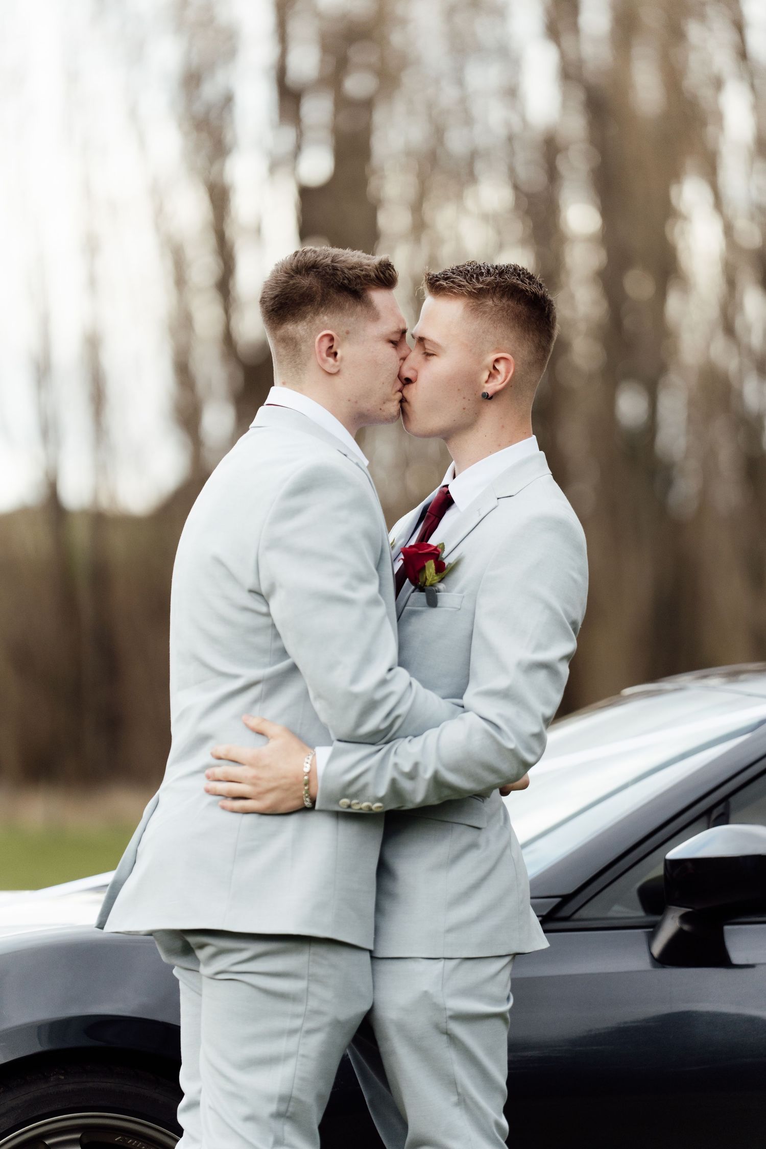 Two grooms in matching light gray suits share a kiss beside a car on their wedding day.
