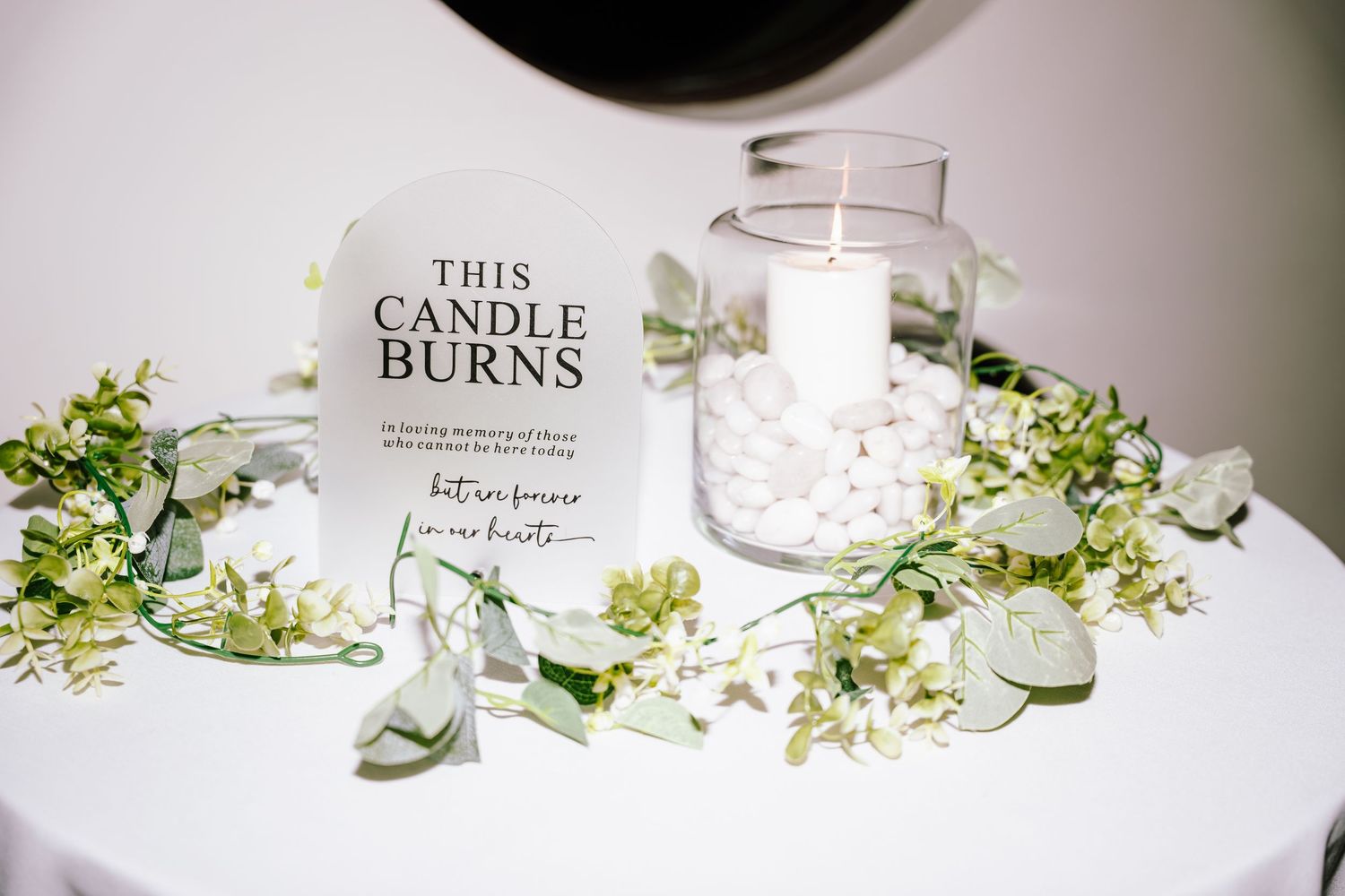 Memorial candle display at wedding with white pillar candle, pebbles, greenery garland, and 
