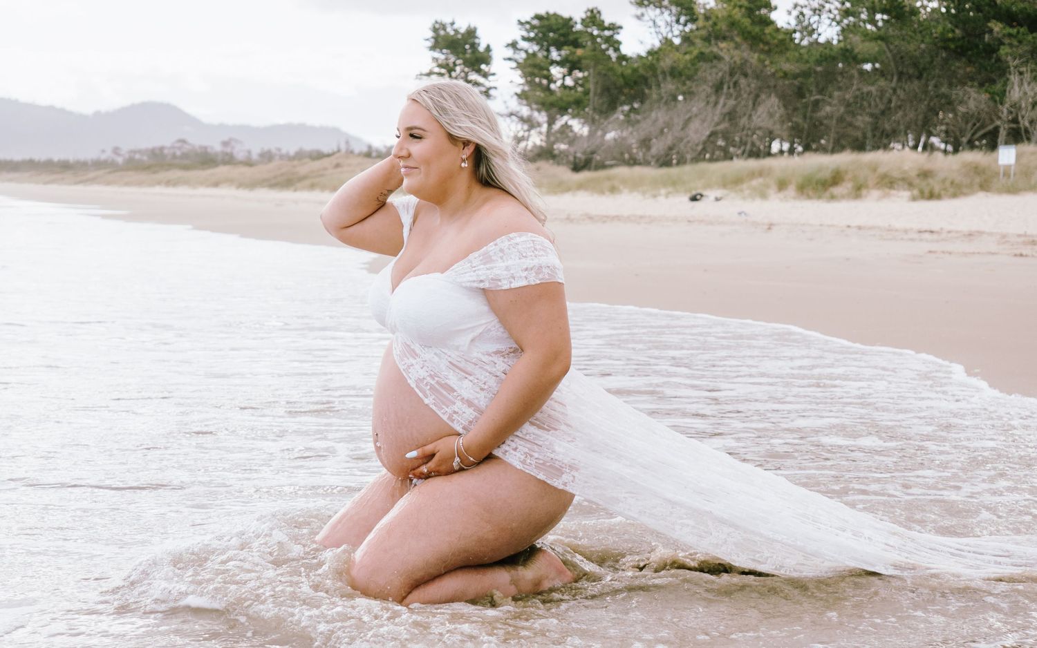 A maternity photoshoot on a sandy beach with soft waves and mountains in the background.