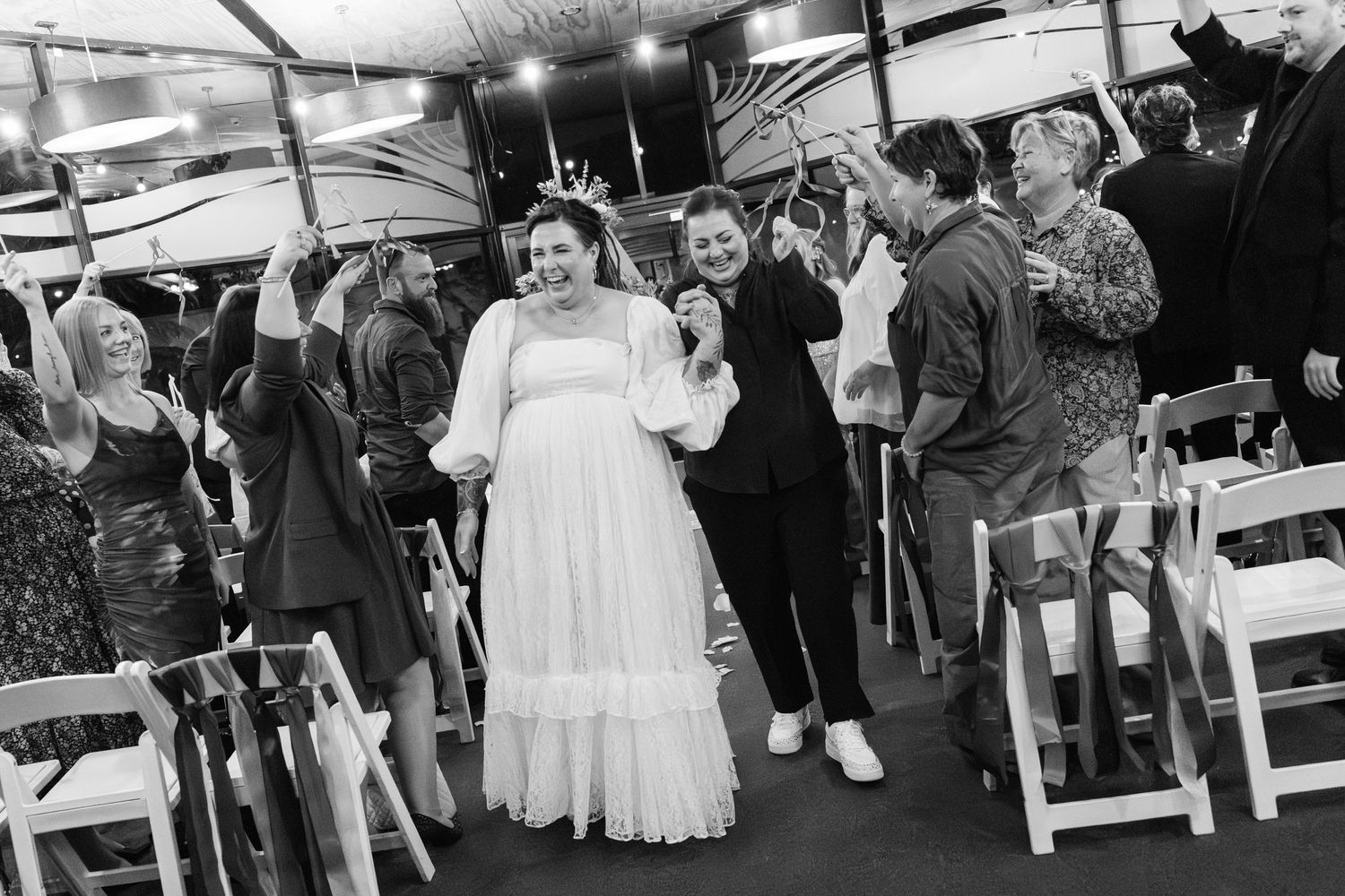 Black and white photo of a bride in white gown walking joyfully through cheering wedding guests under a tent.