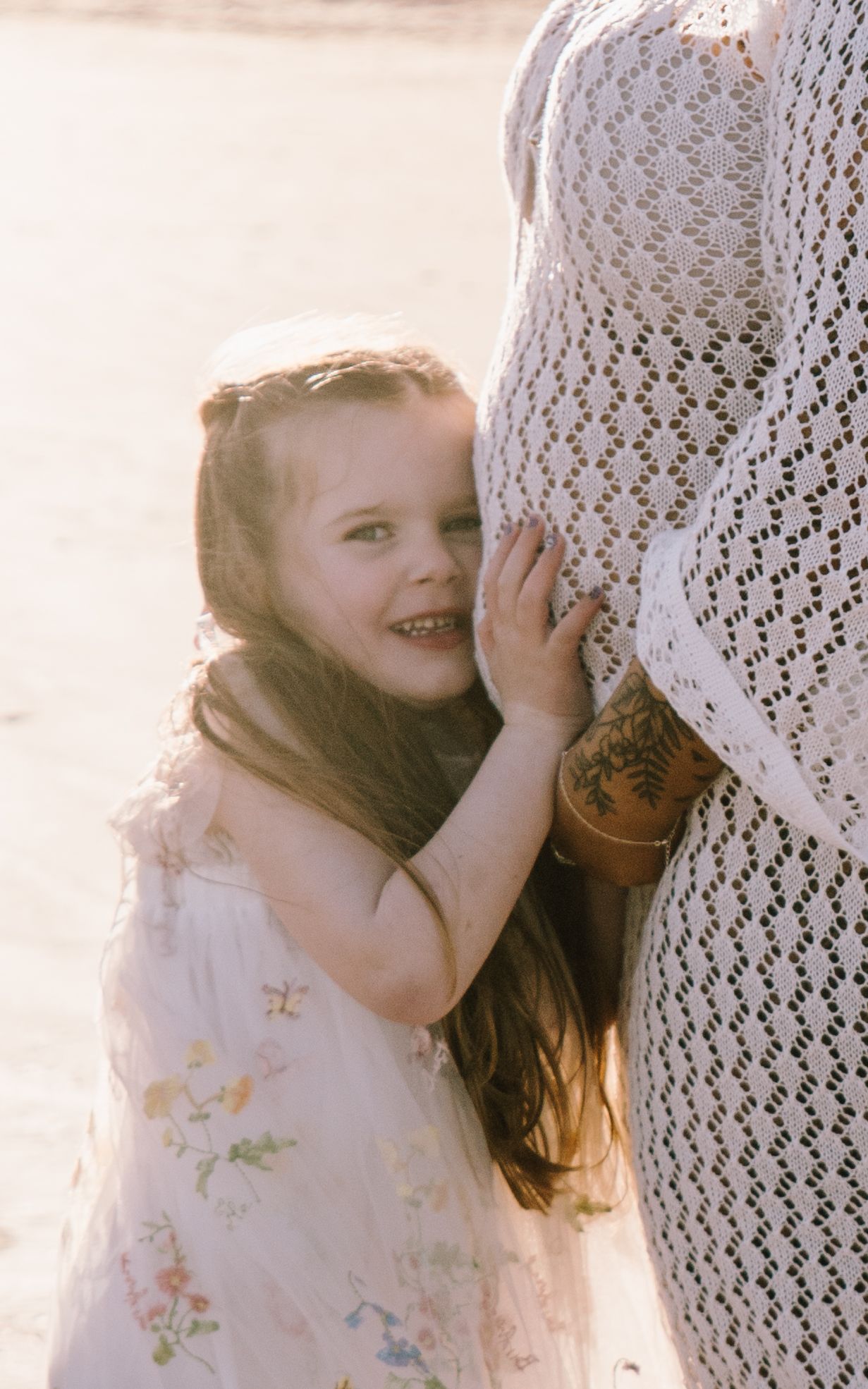A young child in a white dress embraces someone wearing lace in a dreamy, sunlit outdoor setting.