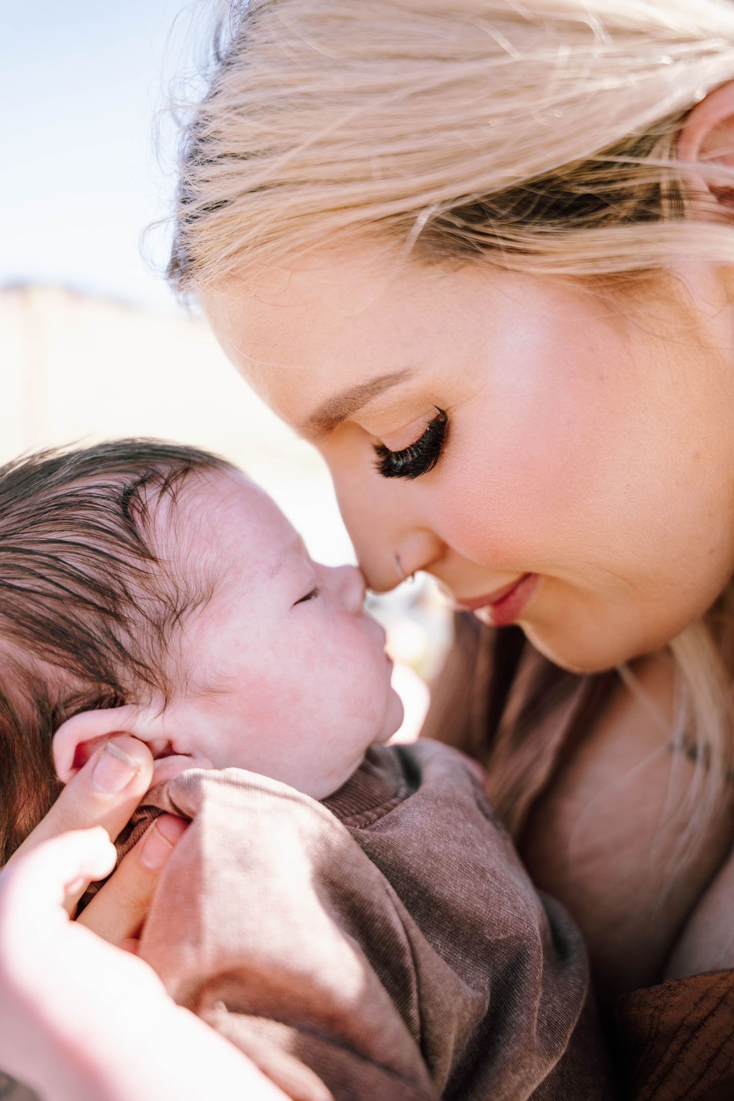 A blonde mother tenderly touches noses with her newborn baby, sharing an intimate bonding moment outdoors.