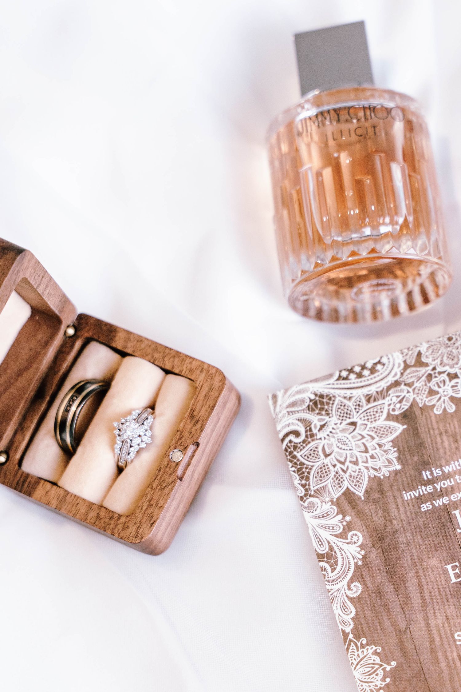Wedding rings in wooden box beside Jimmy Choo perfume bottle and lace invitation on white fabric.