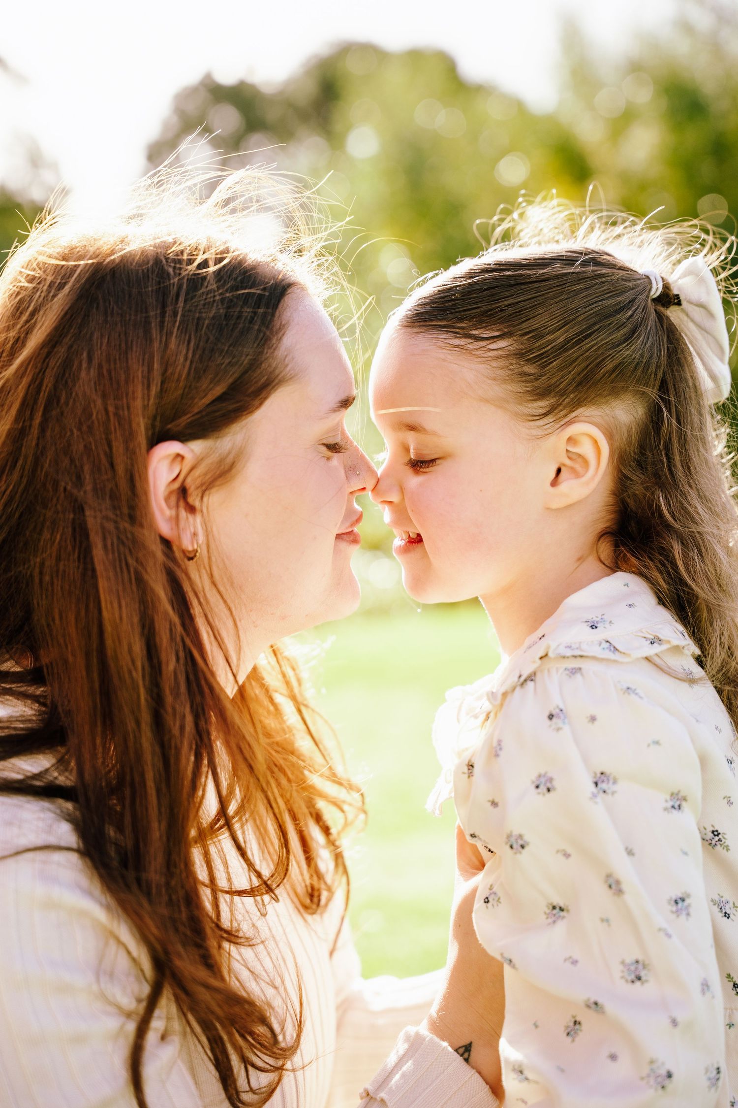 A mother and young daughter share an Eskimo kiss outdoors in warm sunlight, eyes closed in a tender moment.