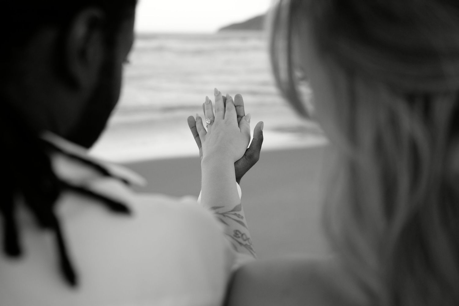 Black and white photo of an interracial couple holding hands at the beach, ocean waves in the background.