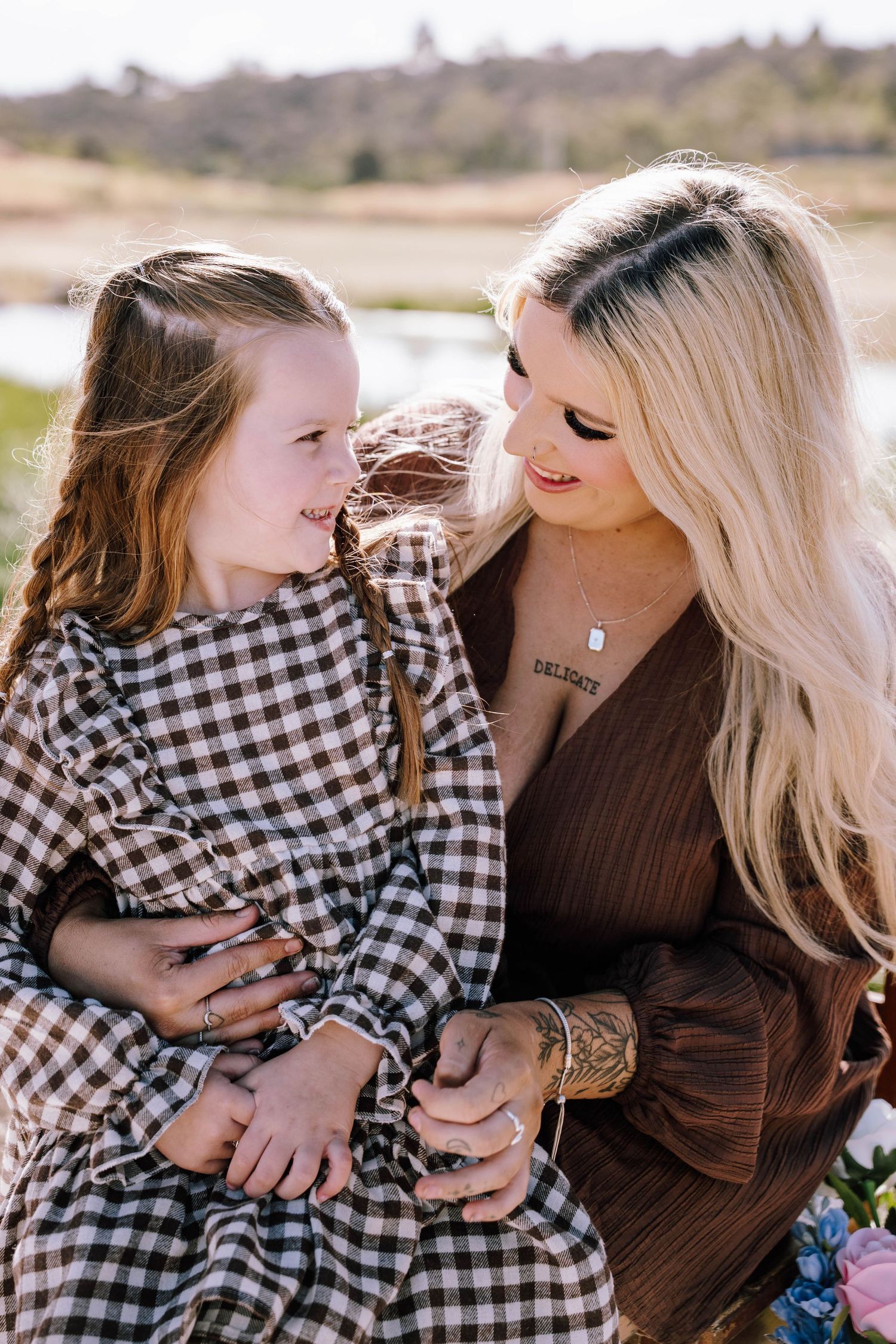 A blonde woman in brown dress holds a young girl in gingham dress, sharing a tender moment outdoors.