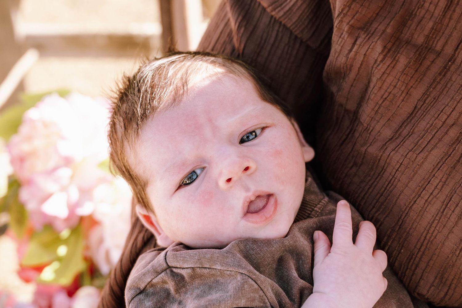 Adorable red-haired newborn baby with blue eyes resting against a caregiver, surrounded by soft pink flowers.