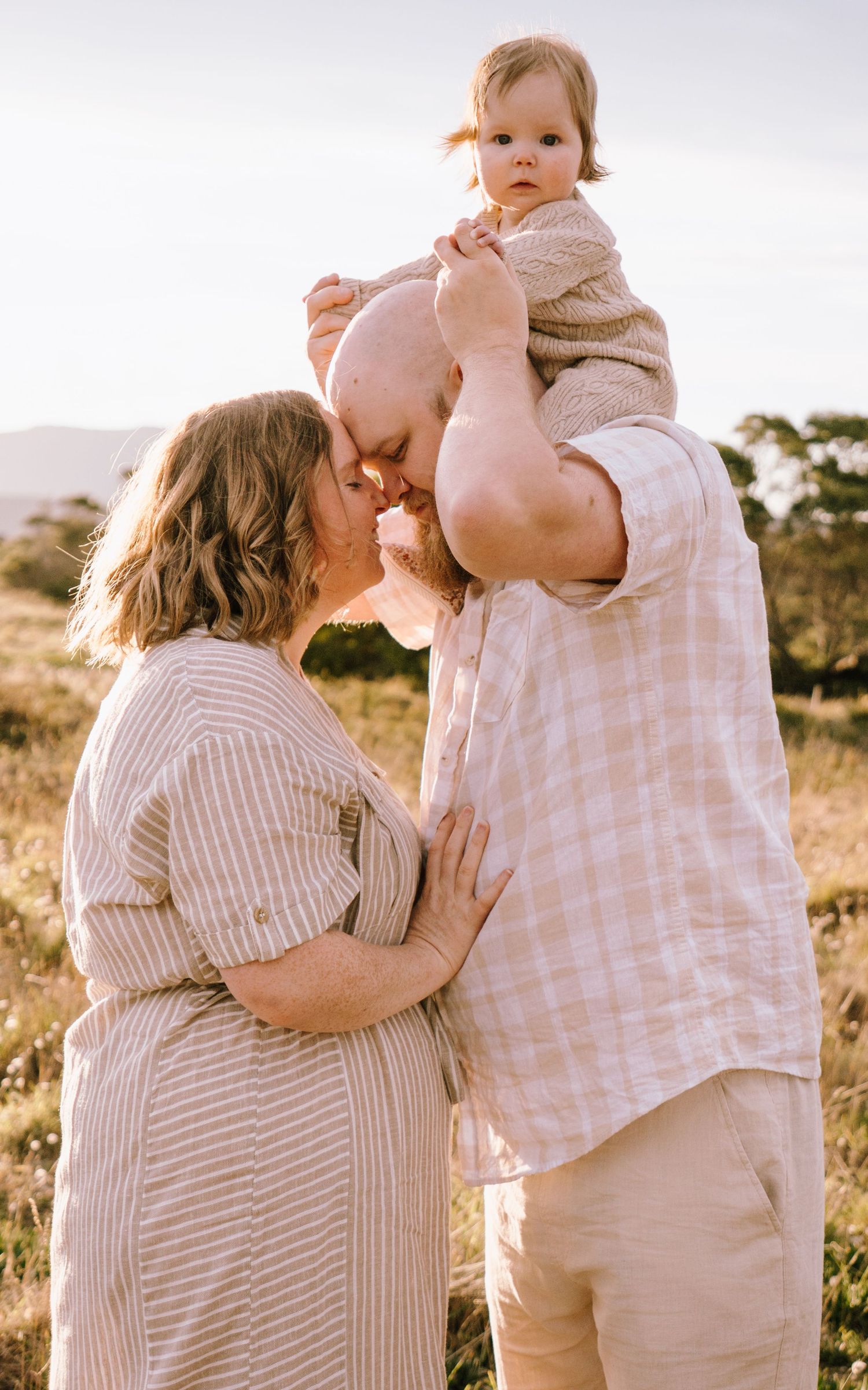 A family shares a tender outdoor moment at sunset in neutral-colored clothing against a natural landscape.