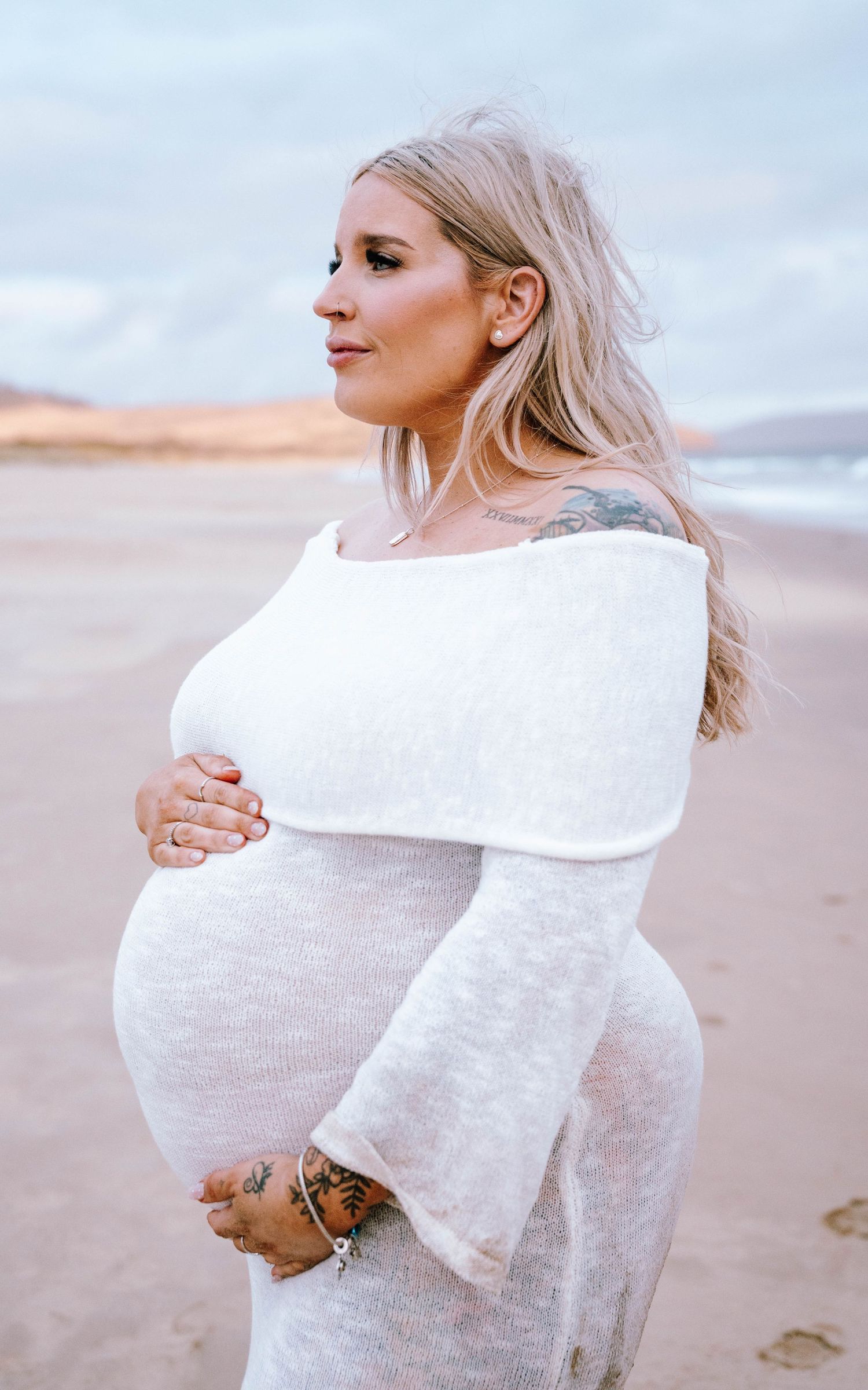A person wearing a white knit sweater poses thoughtfully on a beach during sunset.