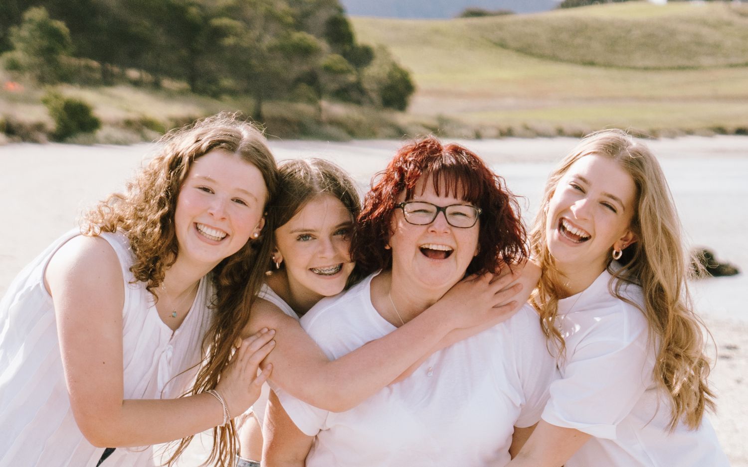 Four smiling women in white tops share a warm embrace outdoors near a beach with green hills behind them.