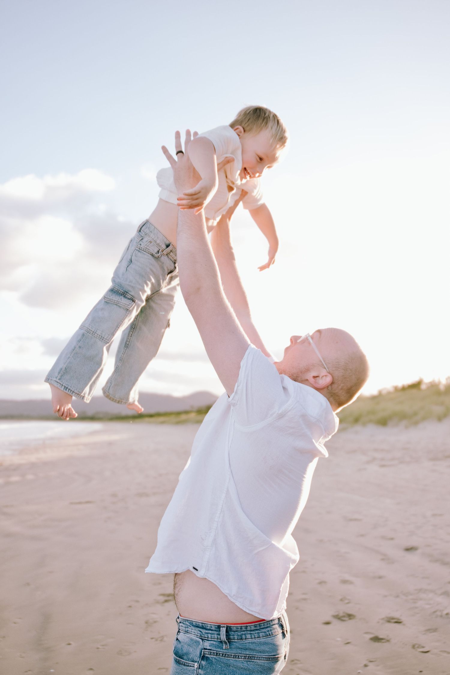 Father joyfully lifting baby high in the air on a sunny sandy beach at golden hour.