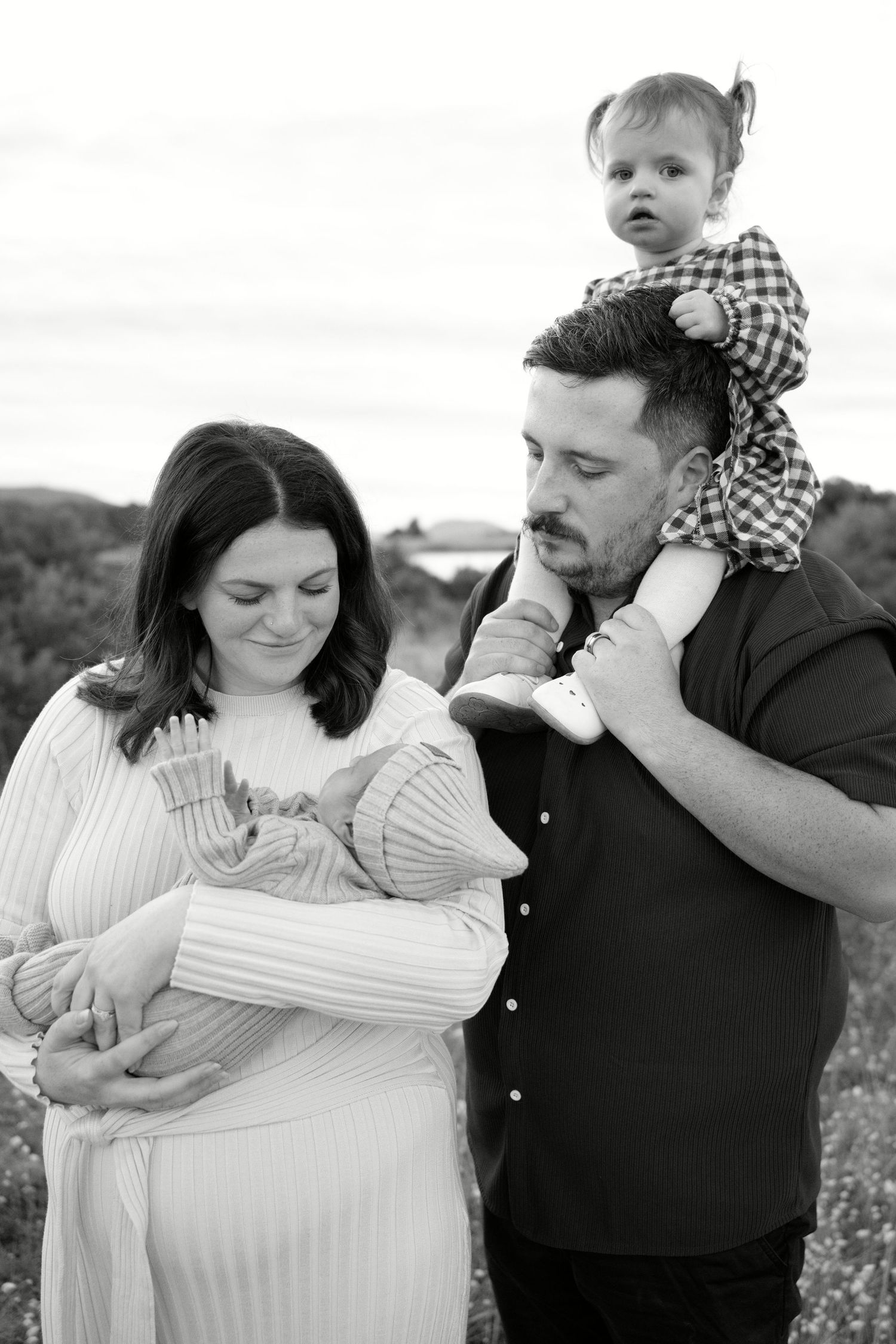 Black and white family portrait of parents with newborn baby and toddler on shoulders outdoors.