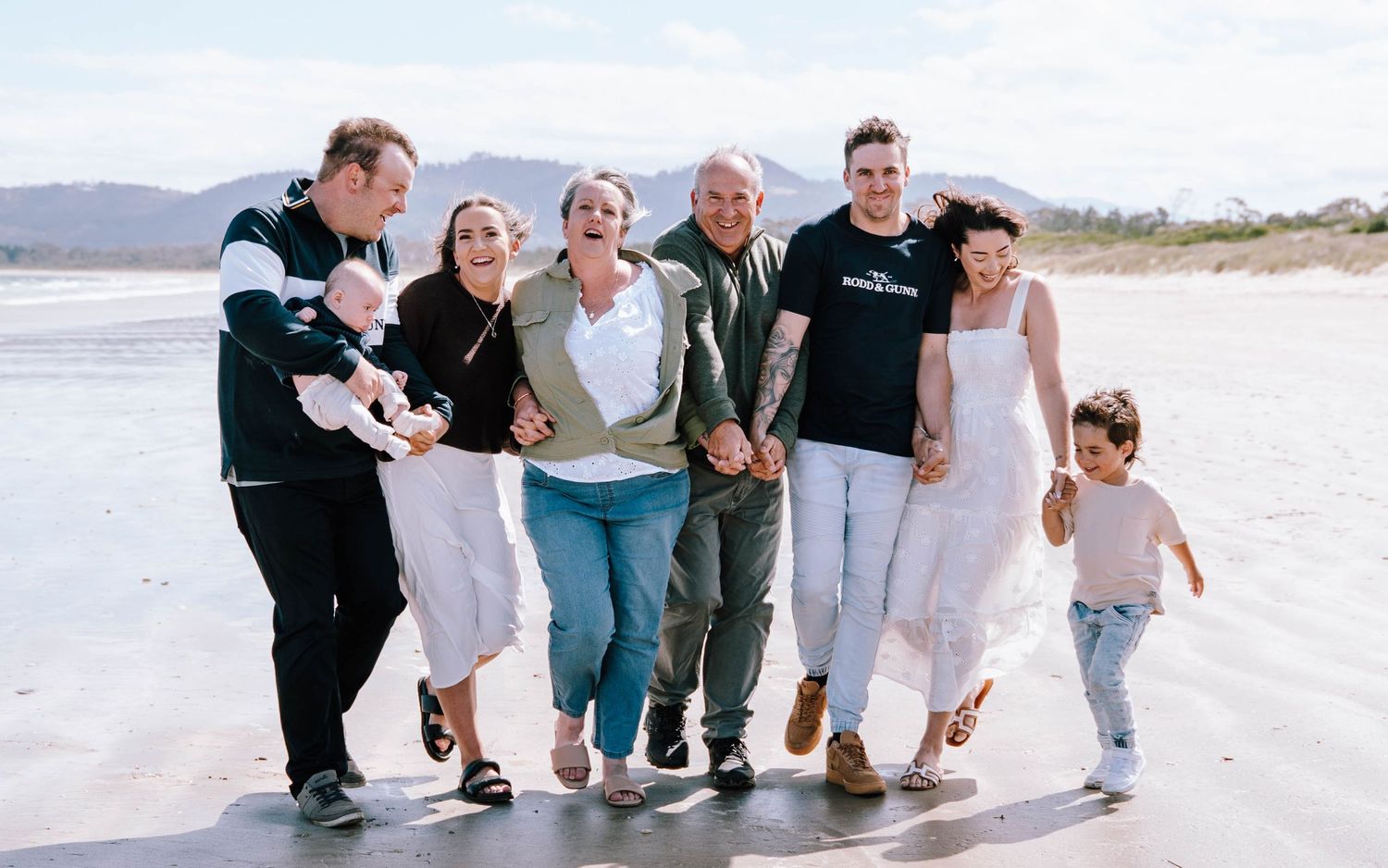 Multi-generational family smiling together on a beach, sharing a joyful moment with mountains in the background.