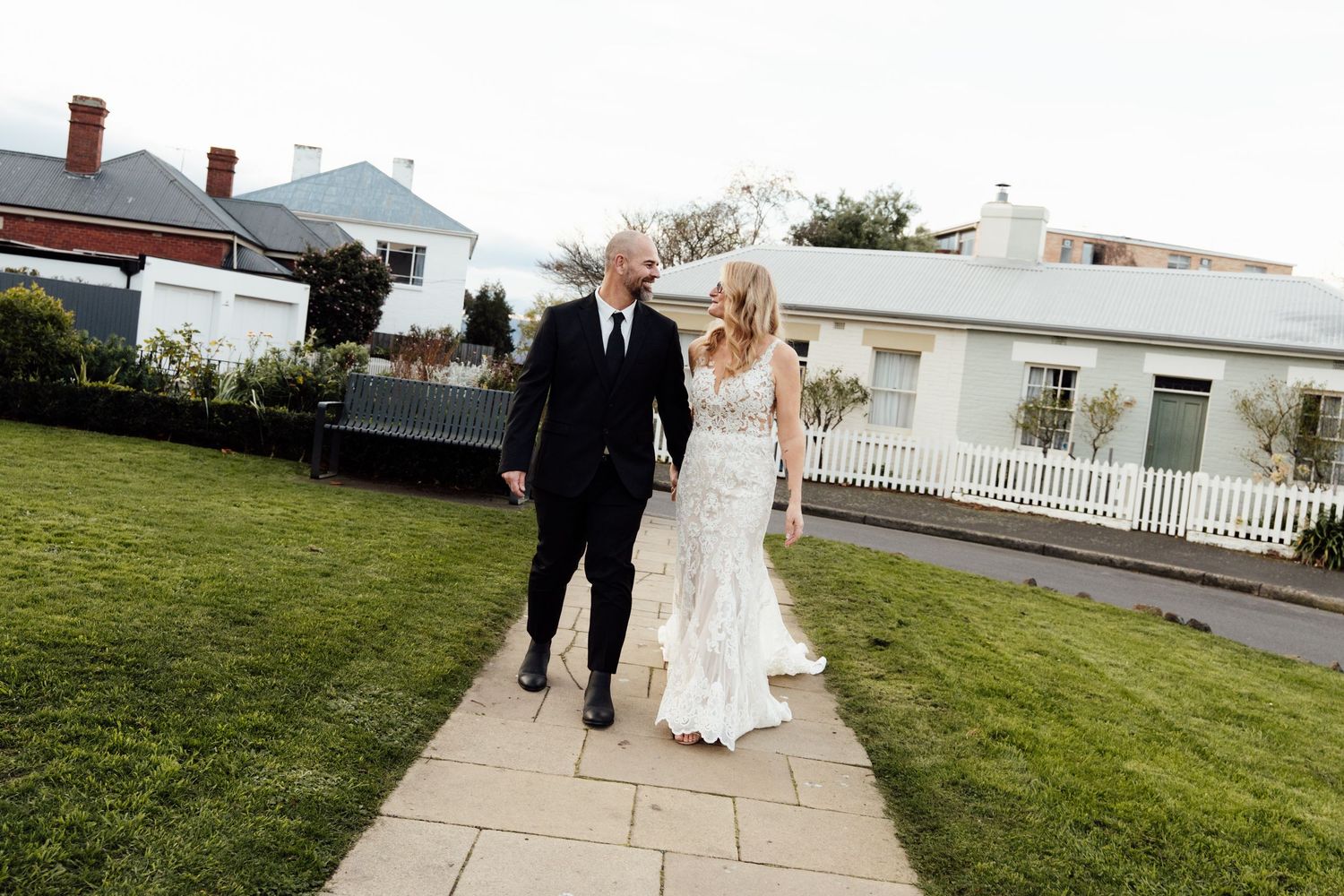 A bride and groom walk hand in hand along a garden path, with white houses and a picket fence in the background.