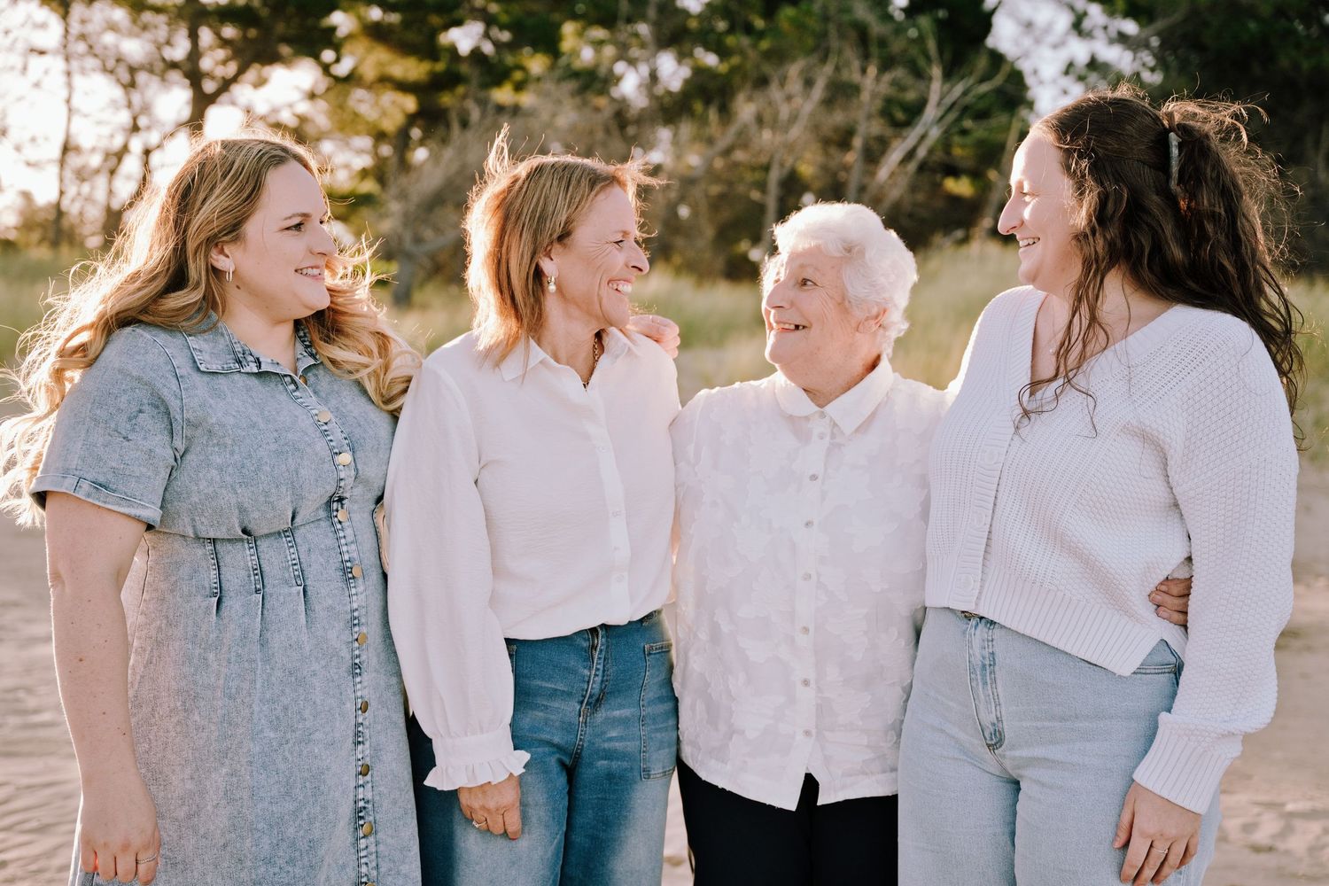 Four generations of women smiling and embracing each other outdoors in a sandy natural setting.