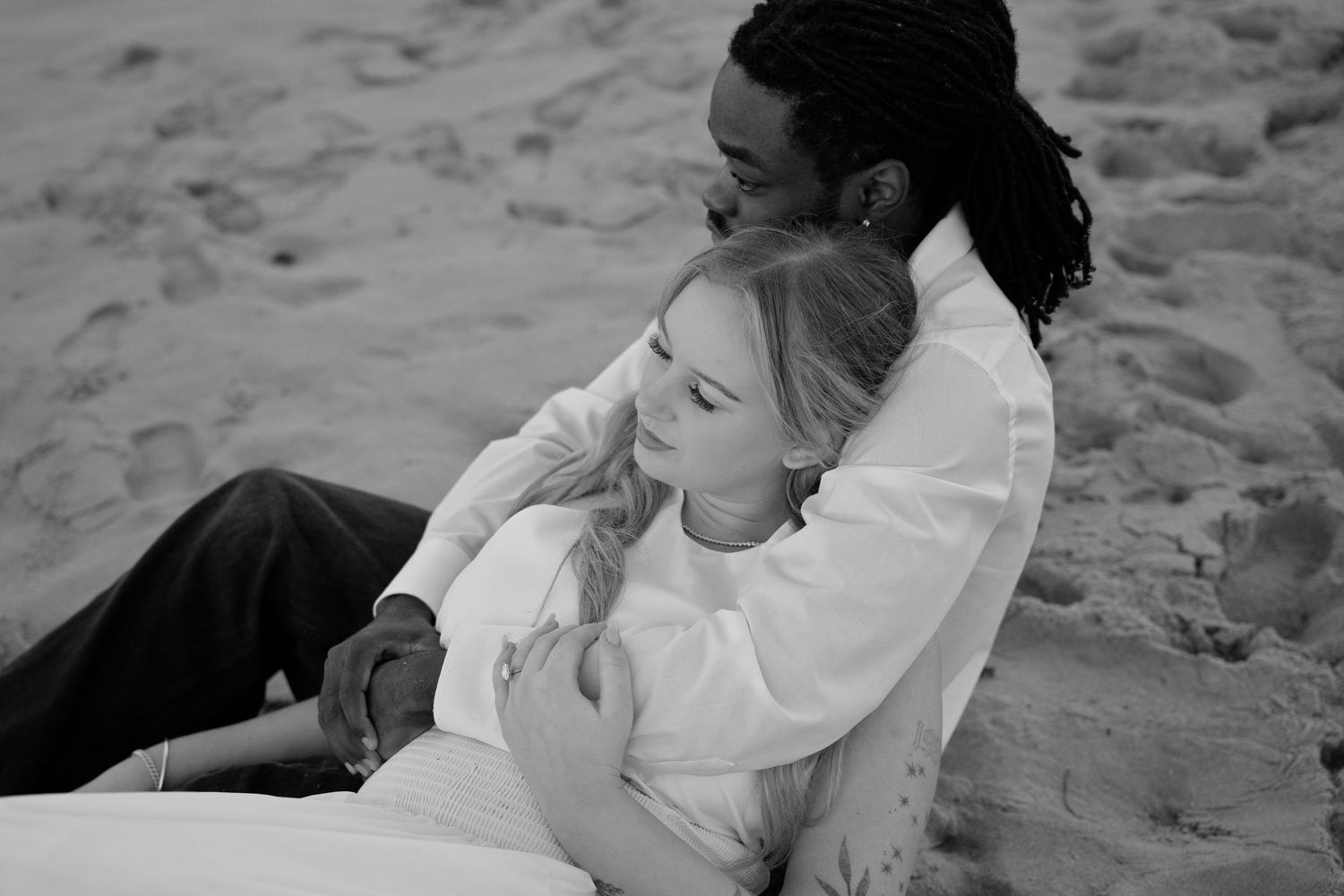 Black and white photo of a couple sitting on sandy beach, woman leaning on mans chest in romantic embrace.
