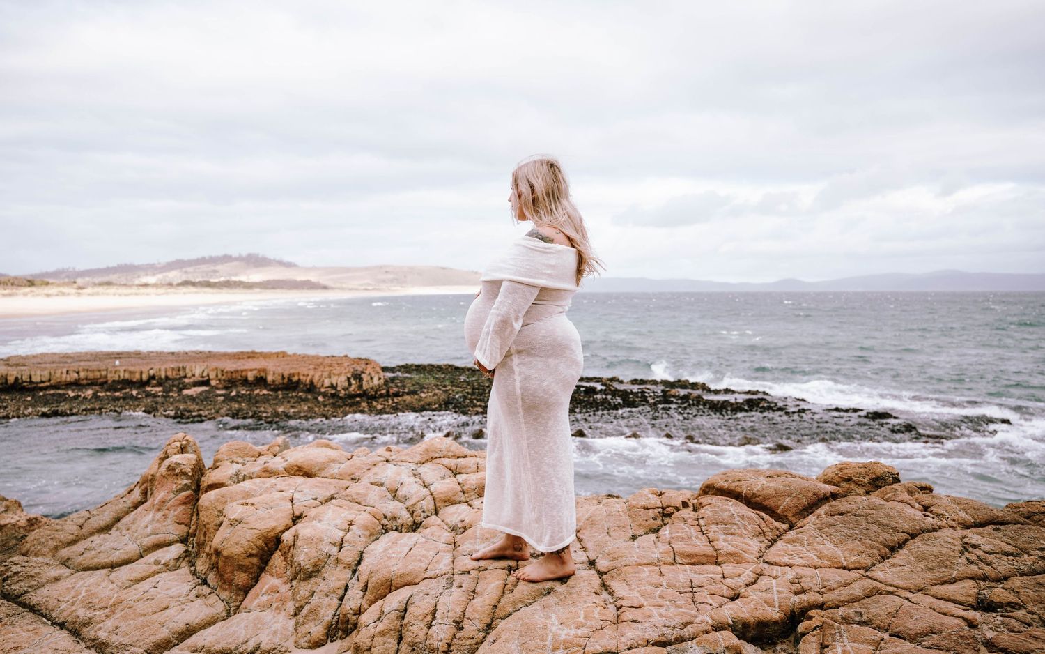 A person in a white dress walks along rugged coastal rocks with ocean waves and cloudy skies in the background.