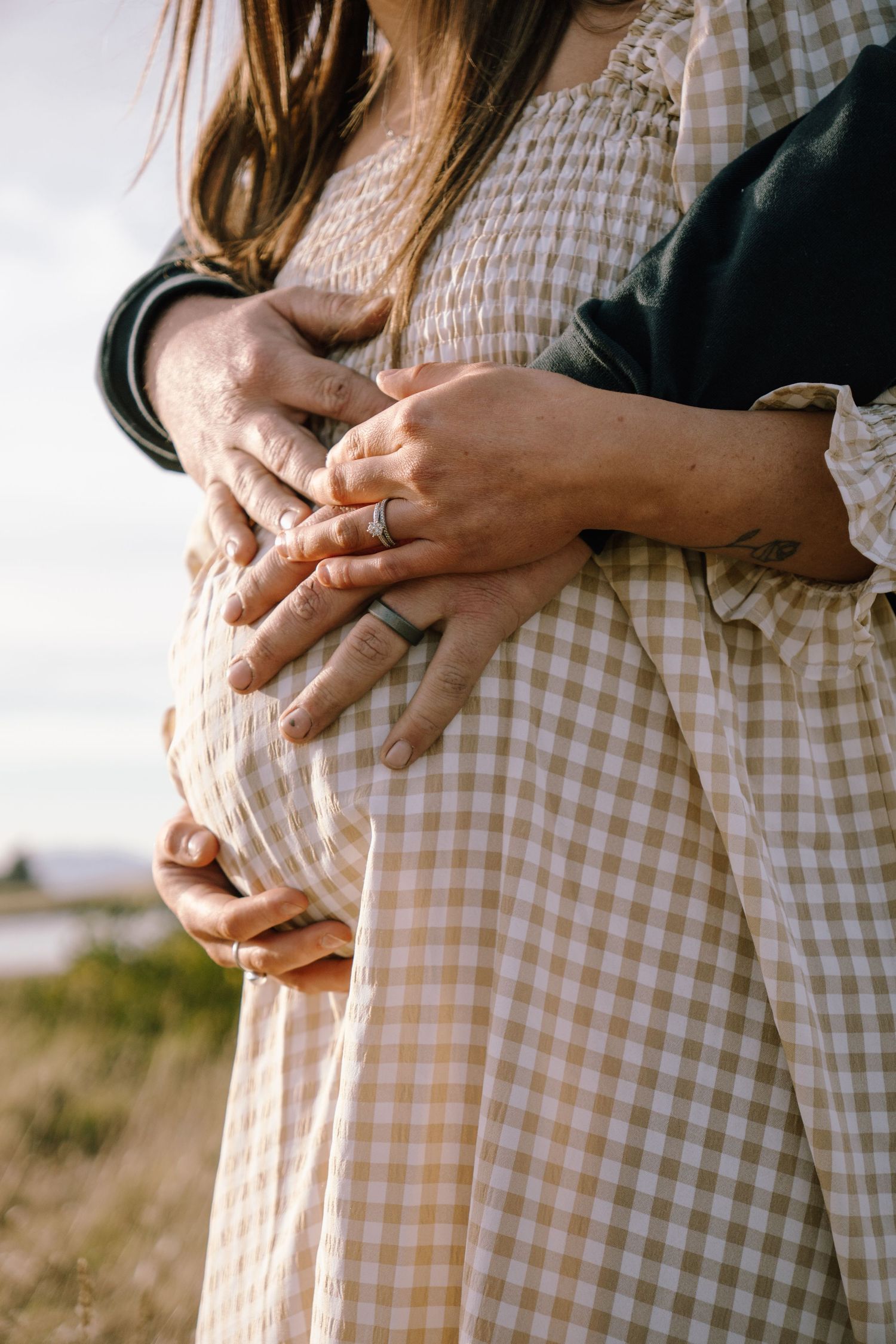 Close-up of hands gently resting on a gingham dress against a sunlit outdoor backdrop.