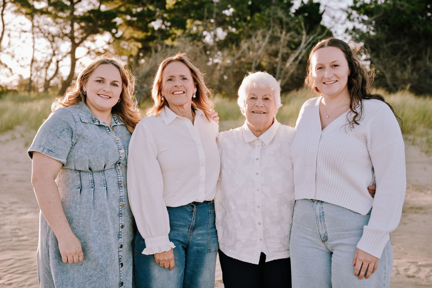 Four women of different generations smile together on a sandy beach at golden hour in casual white and blue outfits.