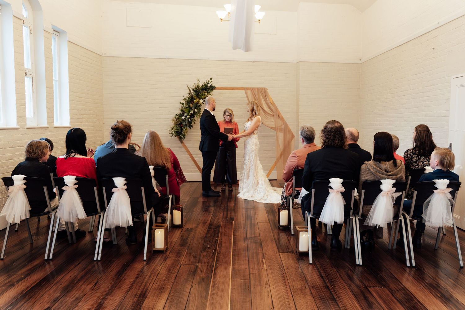Couple exchanging vows at indoor wedding ceremony with wooden arch, seated guests, and white chair decorations.