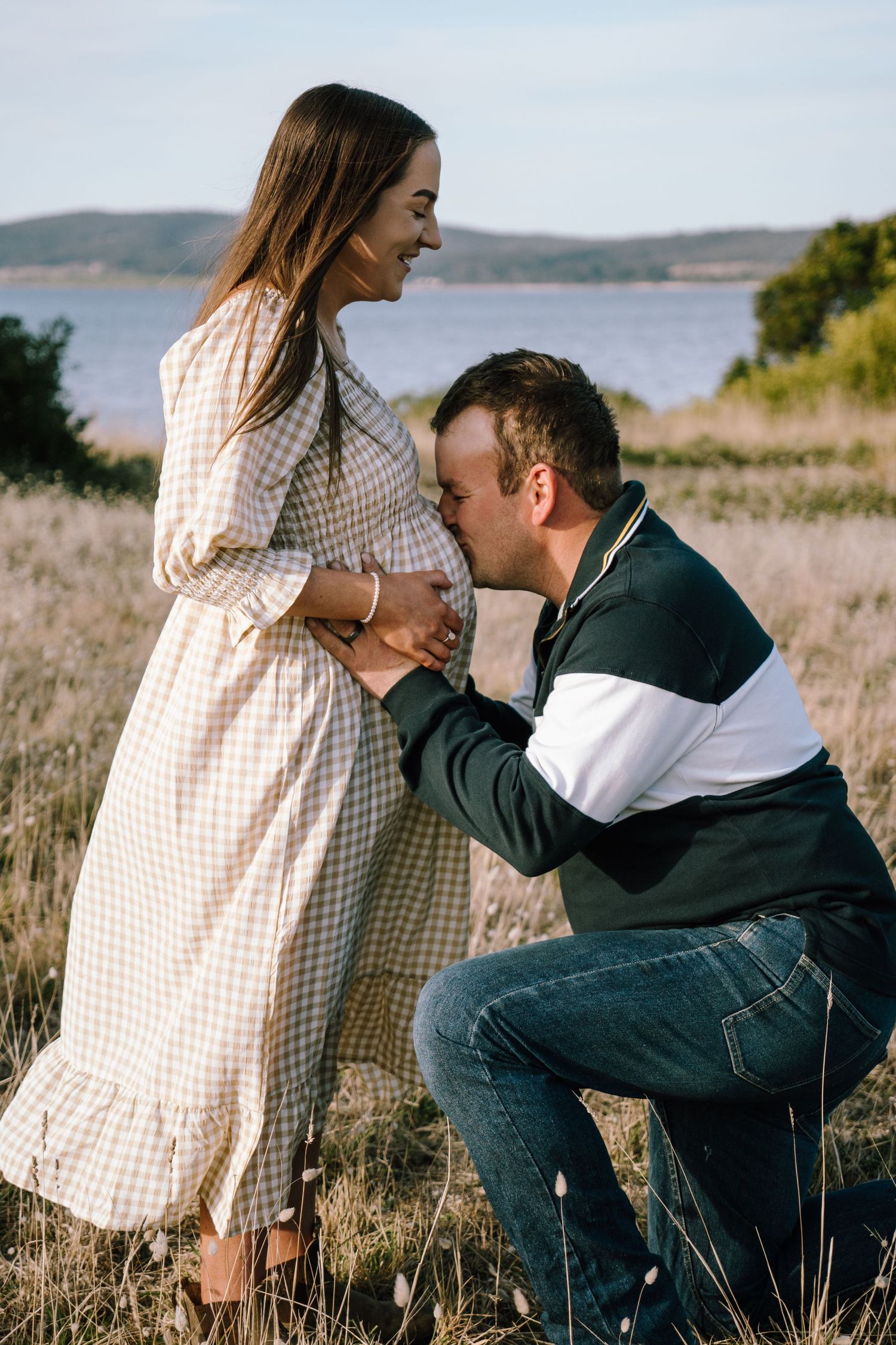 A couple shares tender moments in a grassy field overlooking a scenic lake on a sunny afternoon.