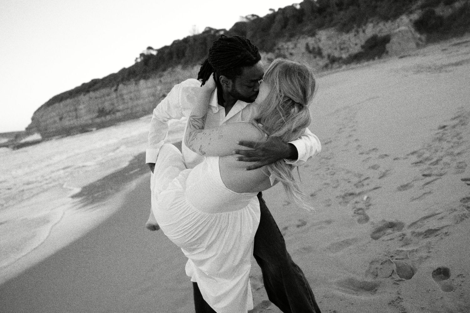 Black and white photo of a couple embracing romantically on a sandy beach with cliffs in the background.