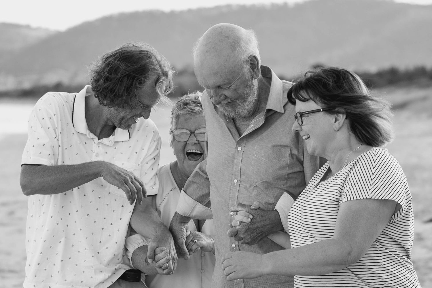 Black and white photo of elderly group laughing together on a beach, sharing a joyful moment outdoors.