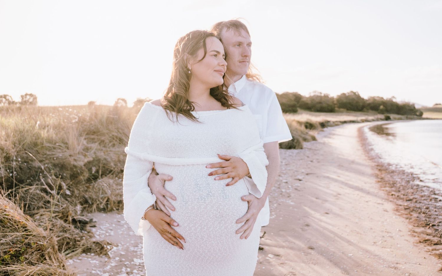 A couple in white outfits embraces on a sunny beach during a romantic maternity photoshoot at sunset.