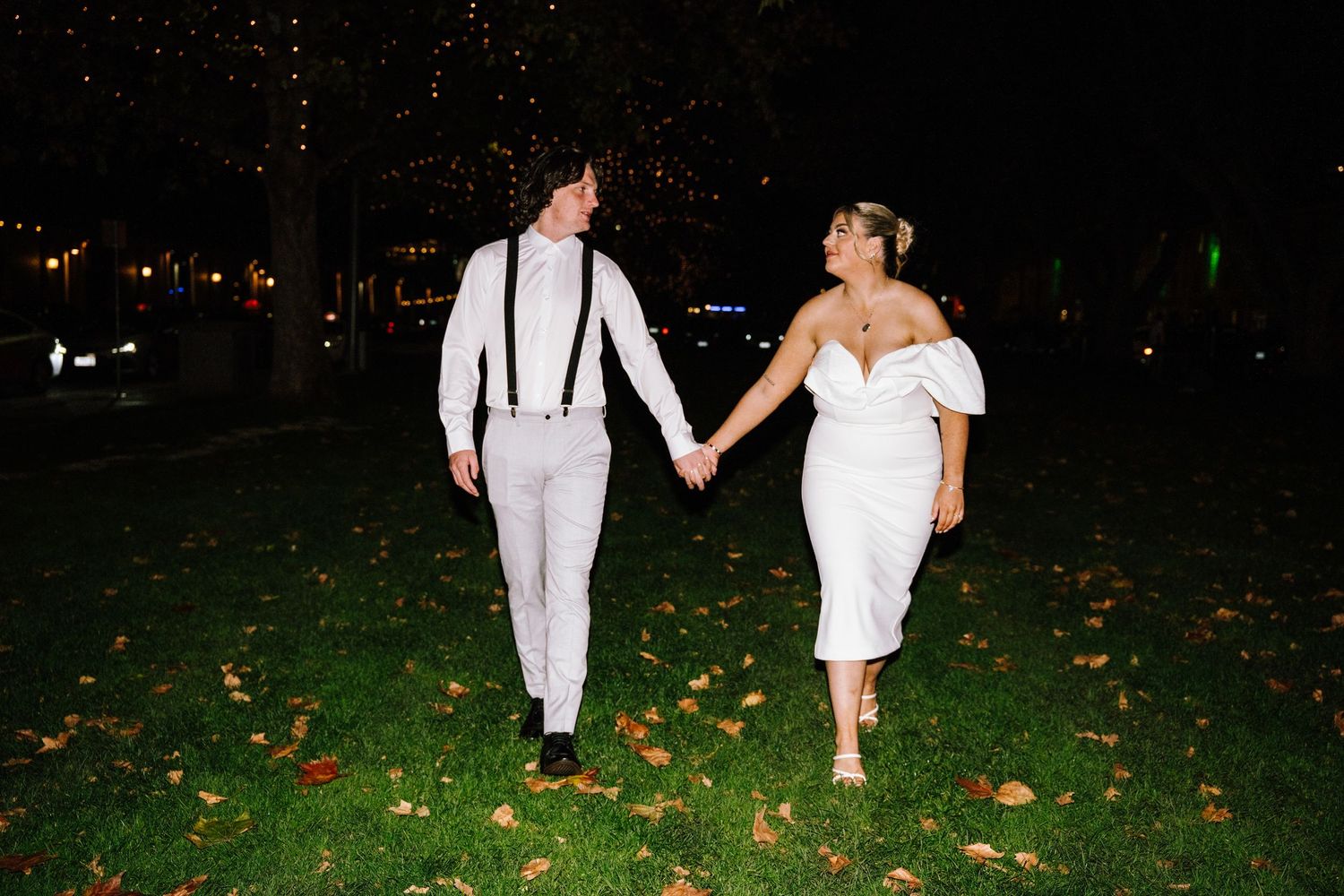 Couple in white wedding attire holding hands walking on green grass at night under twinkling lights.