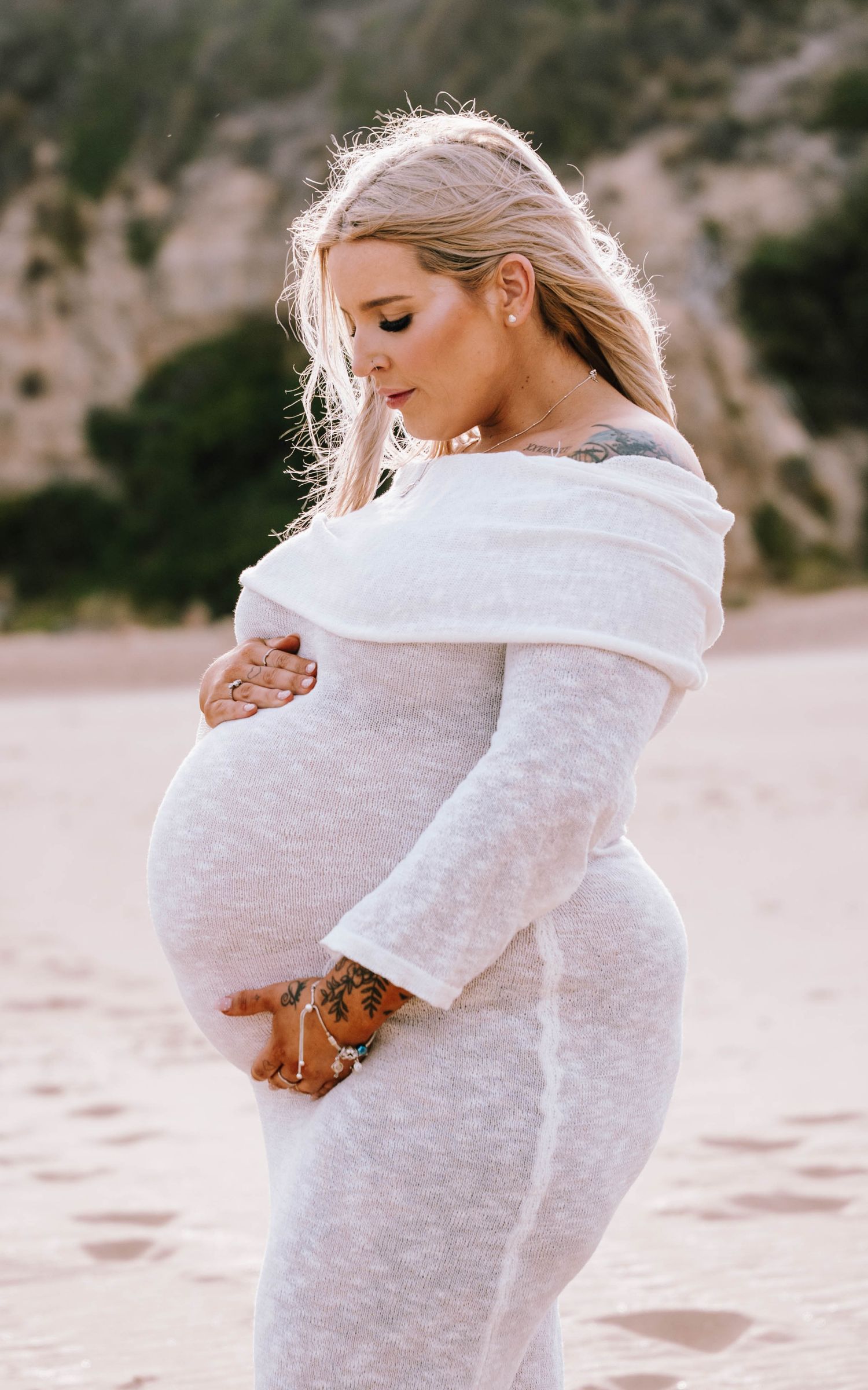 Pregnant woman in white off-shoulder knit dress cradling baby bump on sandy beach with rocky cliffs.