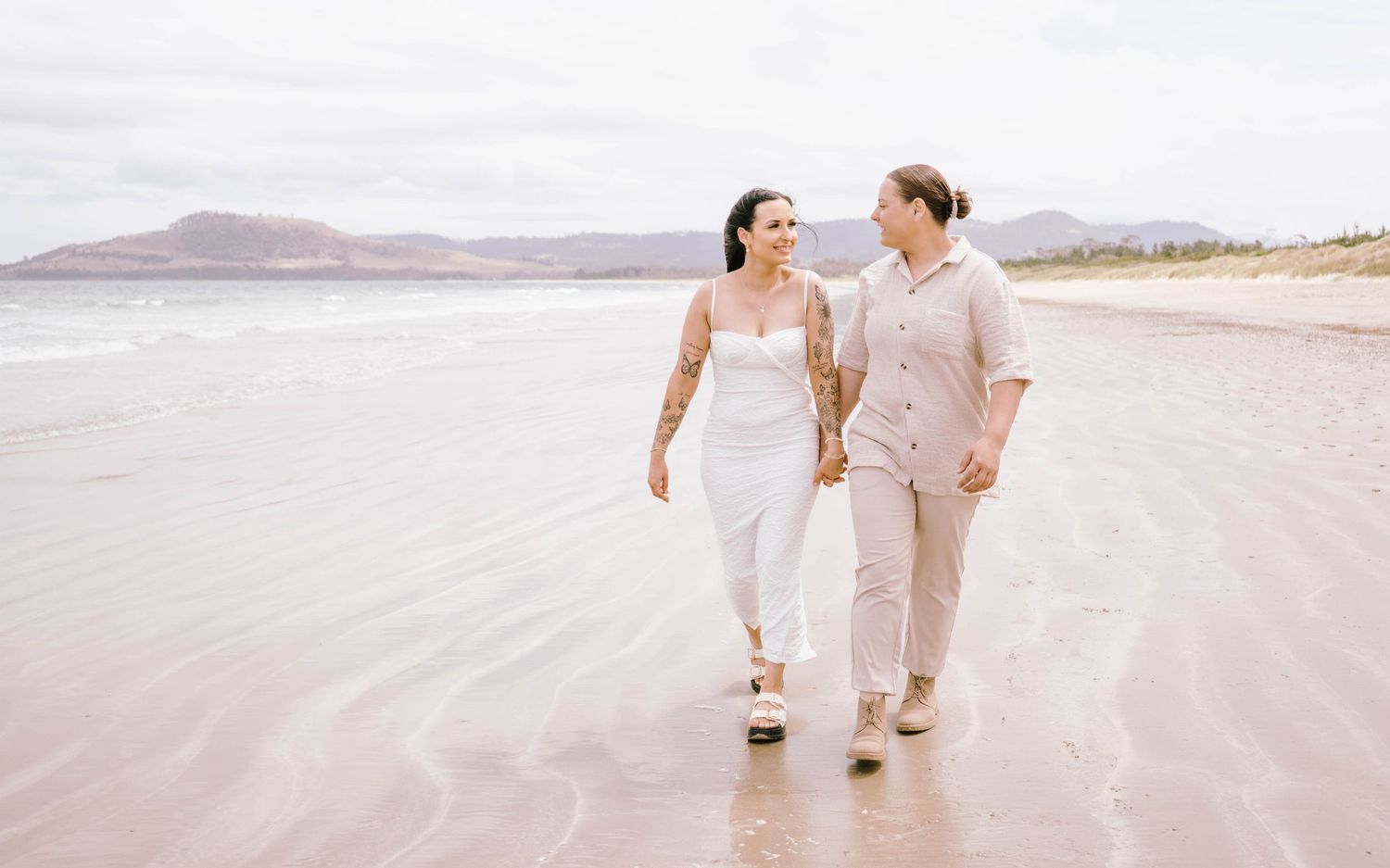 Couple in white and beige outfits walking and playing on a serene beach at sunset.