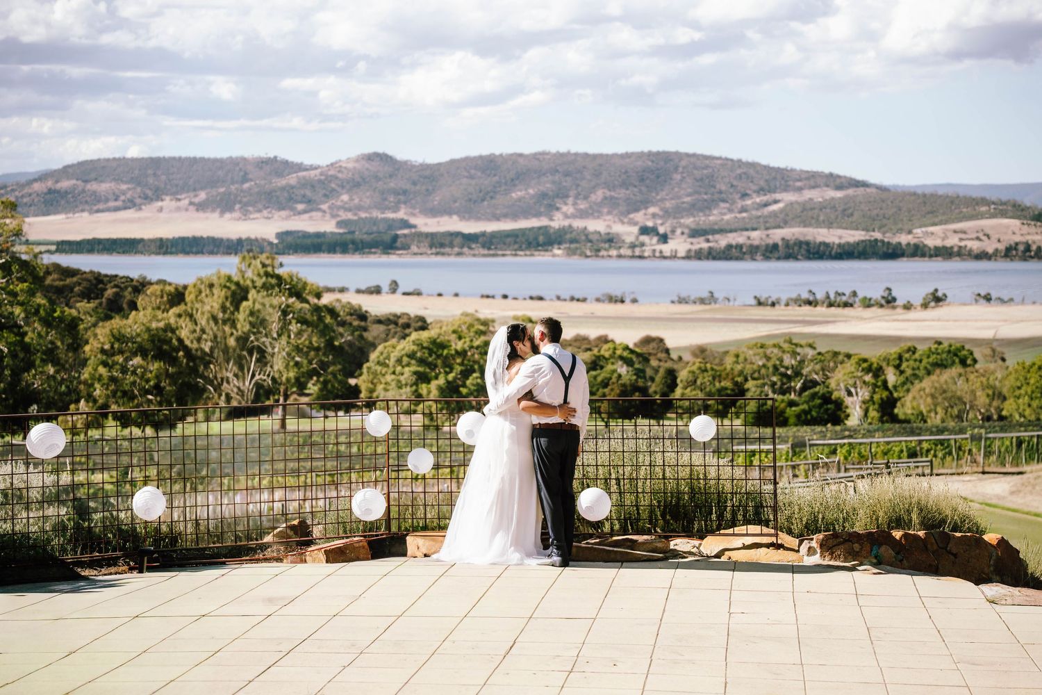 Bride and groom share a romantic kiss overlooking a scenic lake and rolling hills at an outdoor wedding venue.