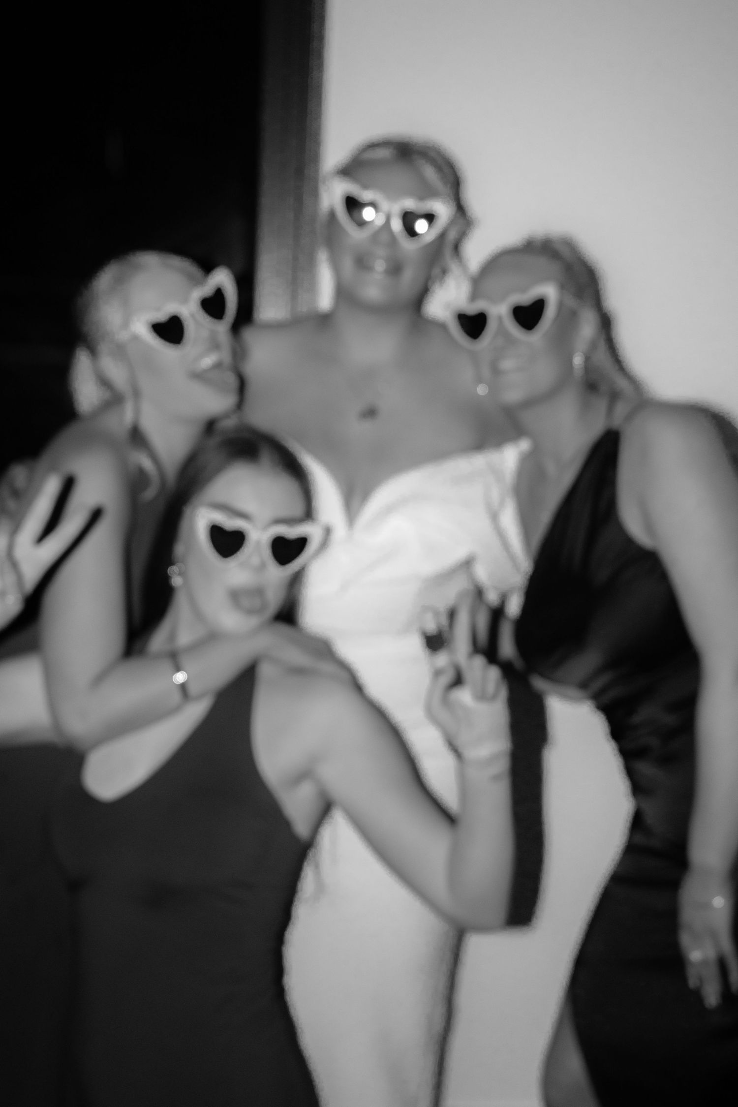 Black and white photo of bride and friends posing in heart-shaped sunglasses at an engagement party.