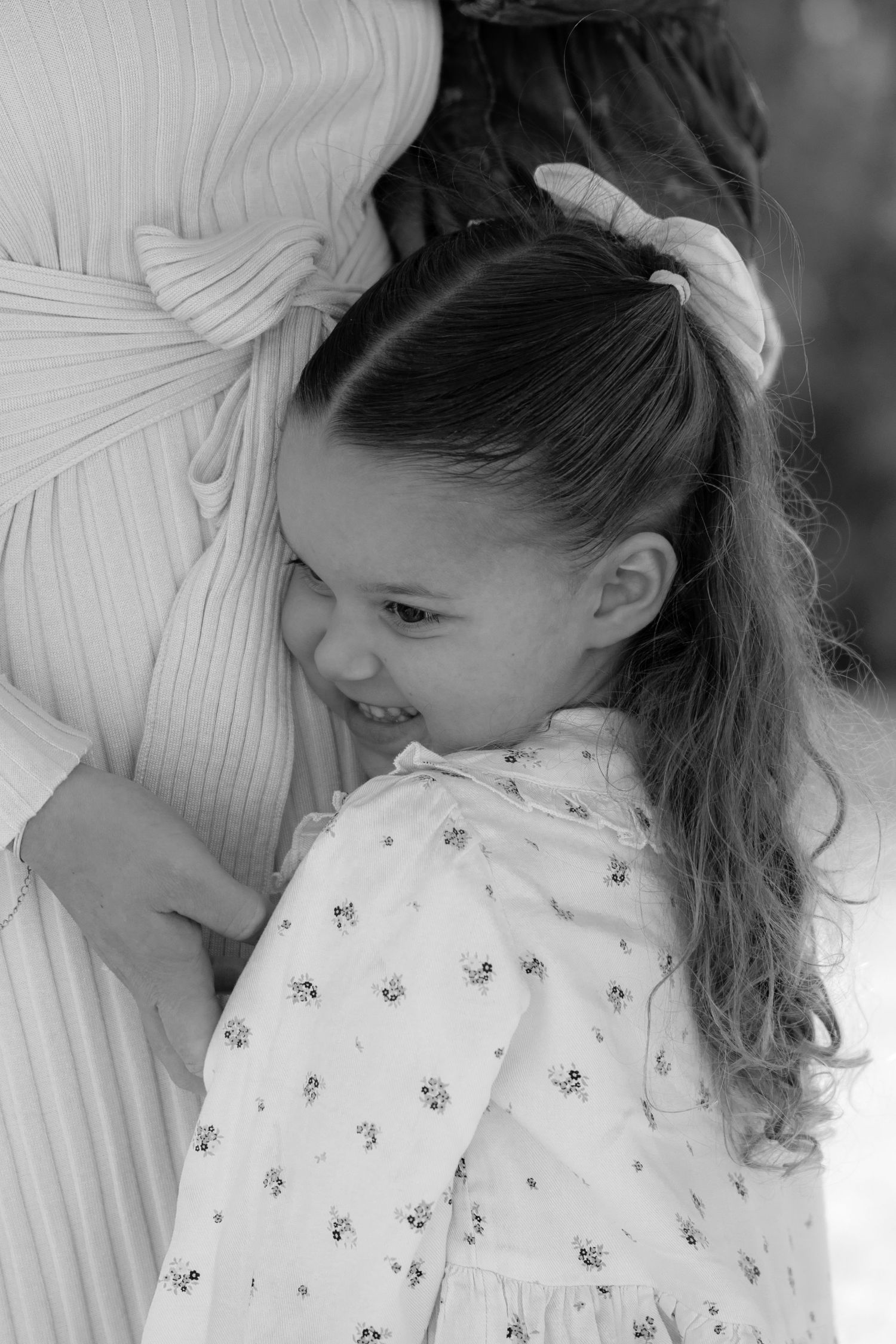 Black and white photo of a smiling young girl hugging an adult, wearing a floral dress with hair in a ponytail.