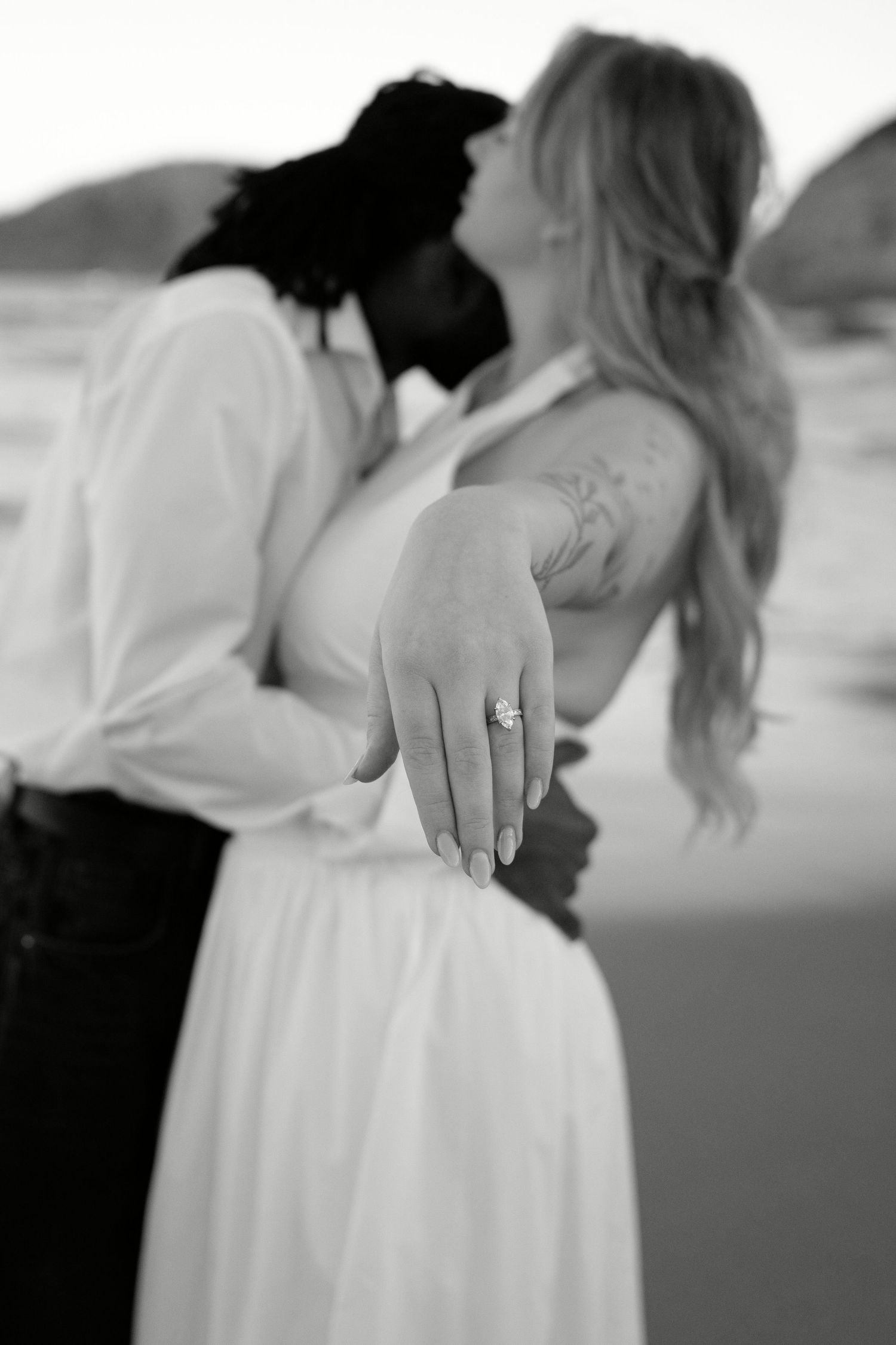 Black and white photo of couple kissing while showing off engagement ring on beach.