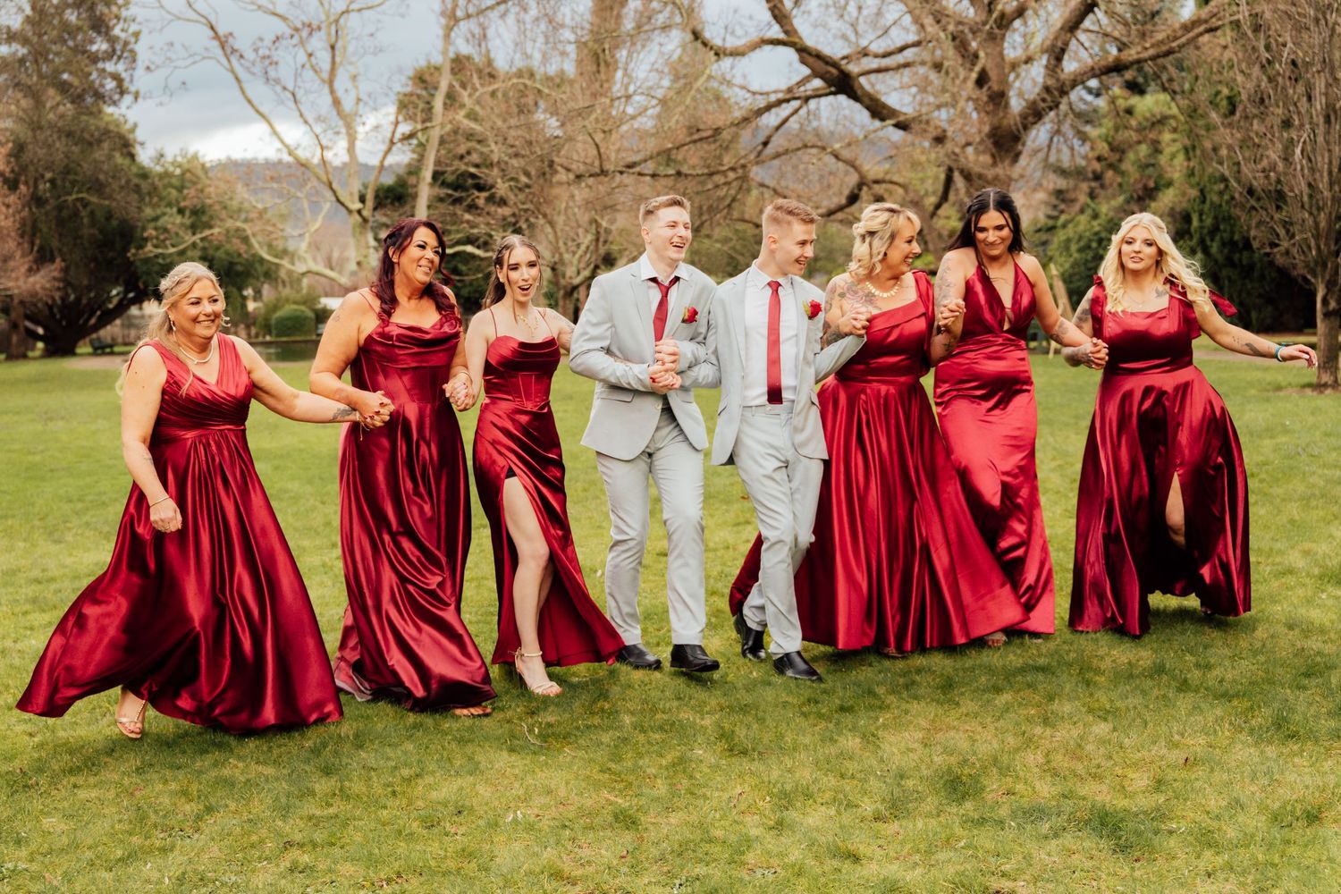 Two grooms in gray suits walk with six bridesmaids in red satin gowns on a green lawn at an outdoor wedding.