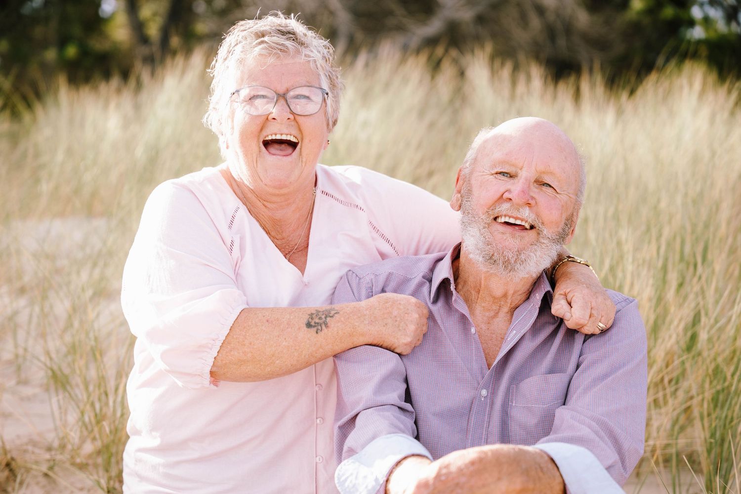 Elderly couple laughing and embracing while sitting together outdoors in a sunny field with tall grass.