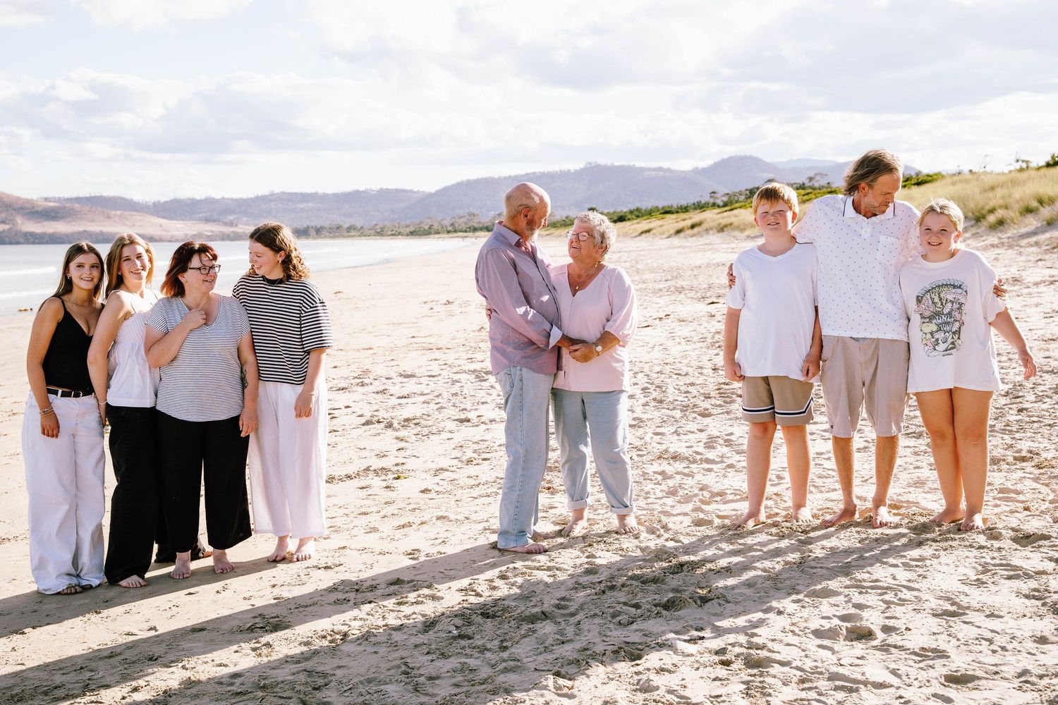 Family group photo on sandy beach with mountains, elderly couple embracing in center, surrounded by children and grandchildren.