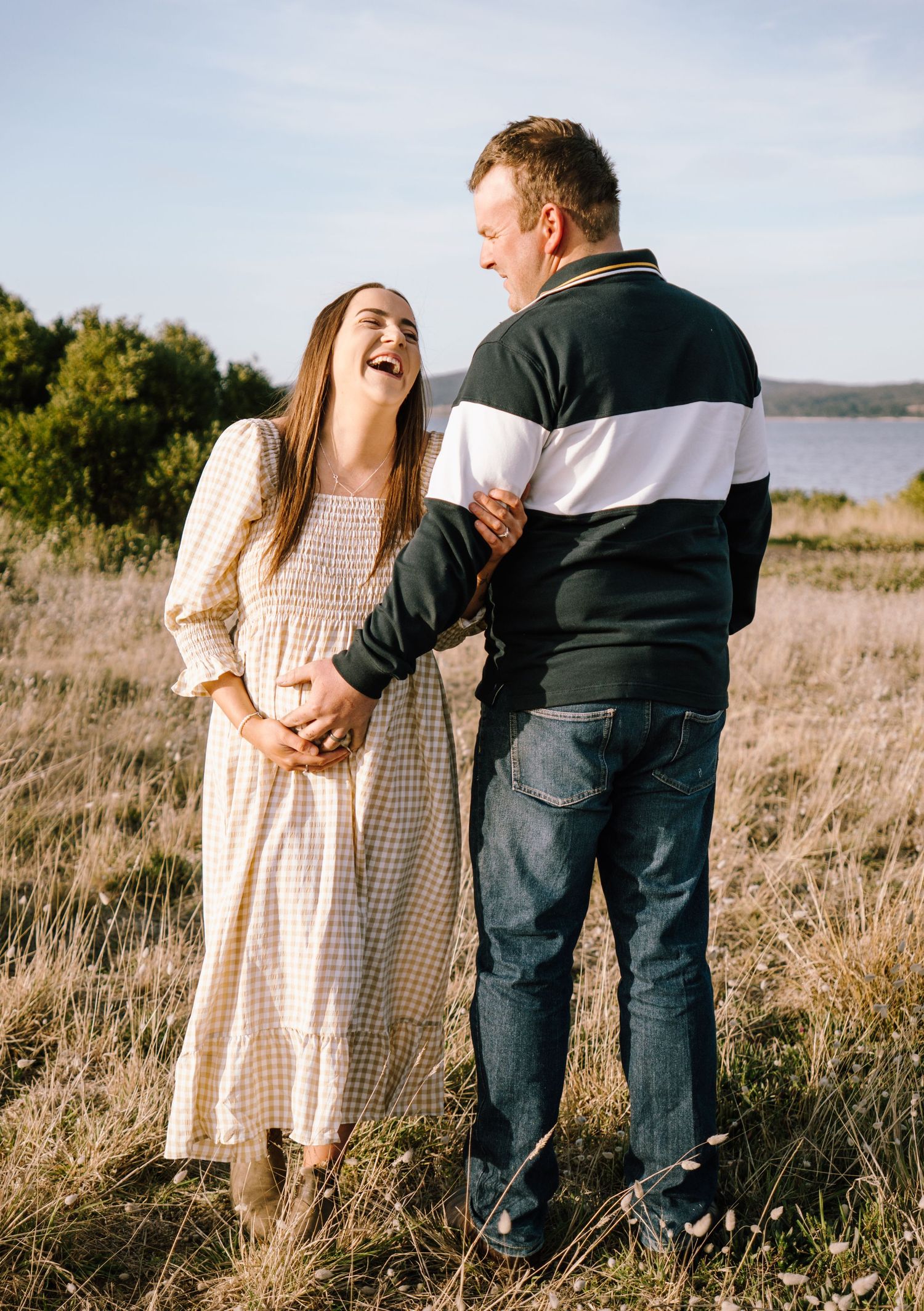 A couple shares a joyful moment together in a field by a lake during golden hour.