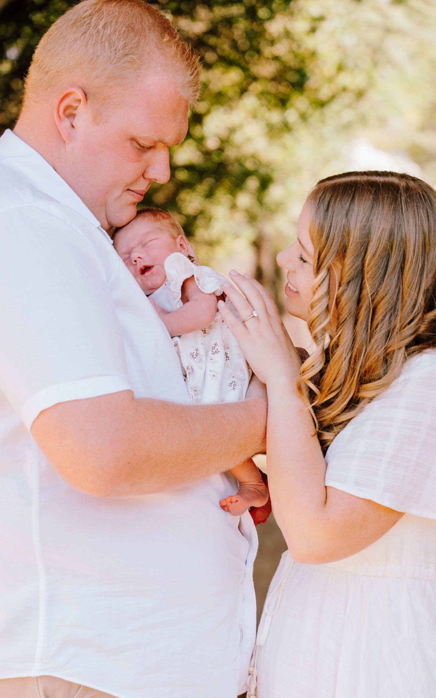 New parents in white outfits tenderly hold their newborn baby outdoors with soft green trees in background.
