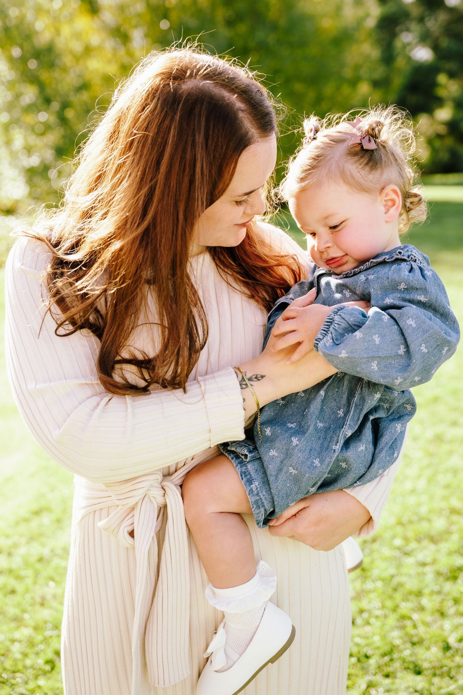 A mother with long red hair holds her toddler daughter in a sunny green park, both wearing casual outfits.