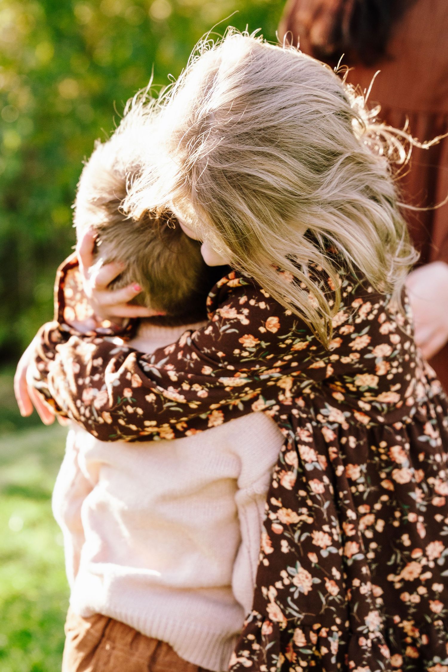 Two children hugging outdoors, one wearing a floral brown dress and the other in a white sweater, surrounded by greenery.