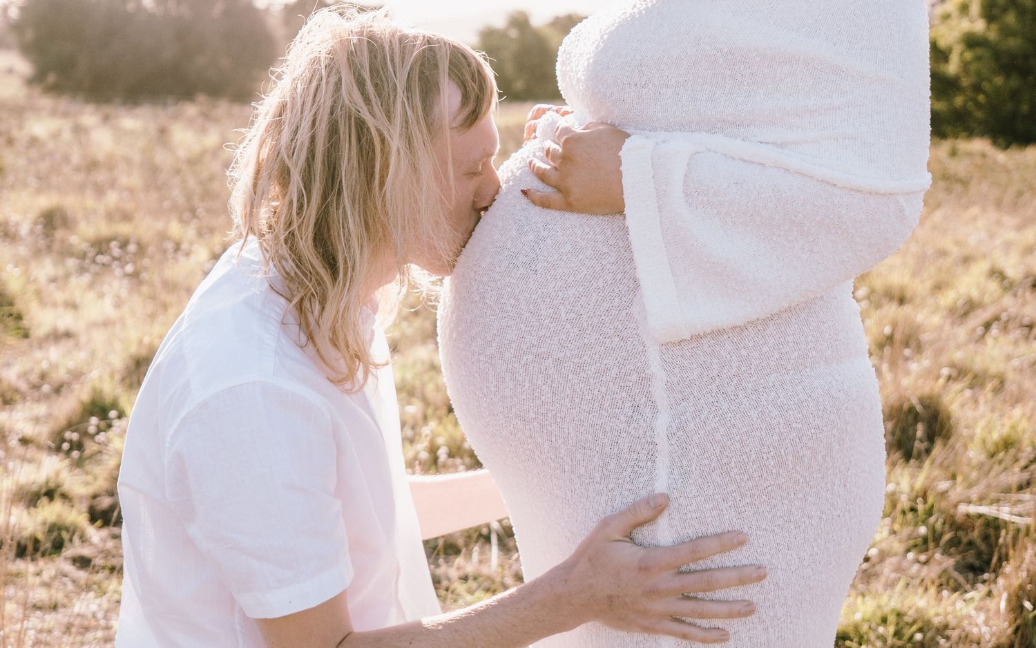A tender moment captured in sunlit field as a person kisses a pregnant belly in white clothing.