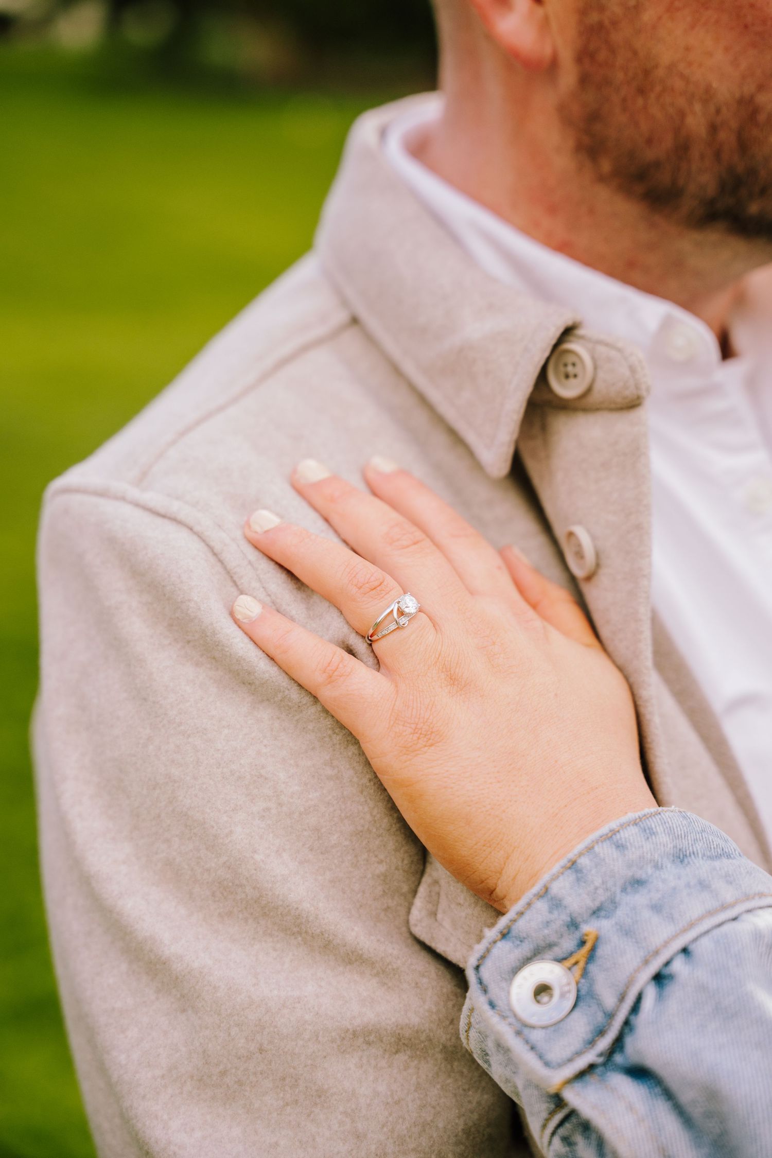 Close-up of a woman's hand with an engagement ring resting on a man's beige coat shoulder.