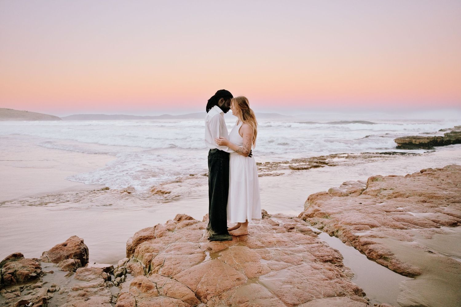 Couple embracing on rocky beach at sunset with pink sky and ocean waves in background.